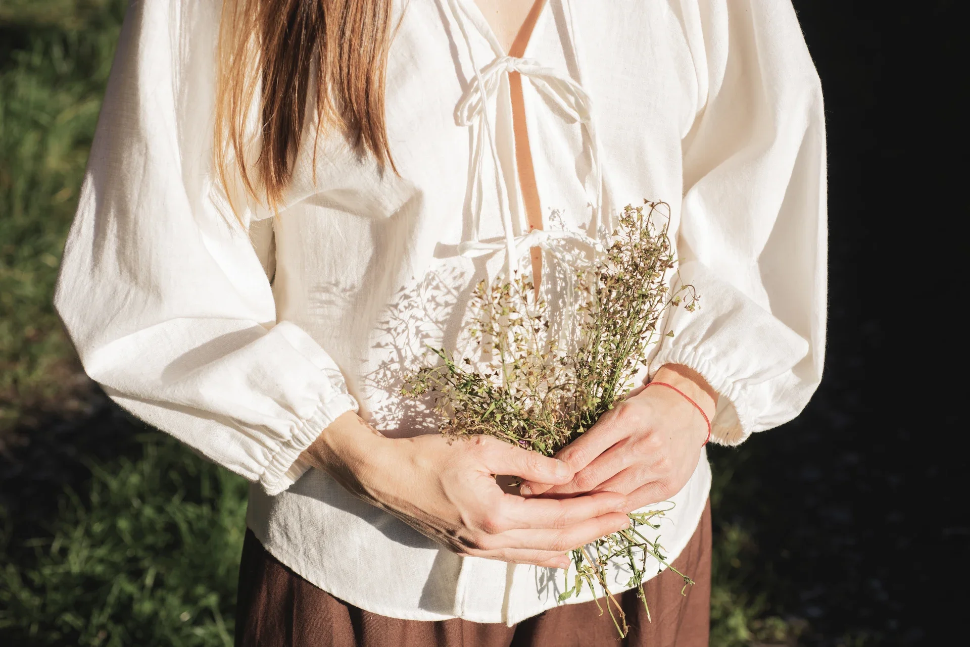 Mujer vistiendo la Blusa Cala blanca de algodón orgánico con lazos delanteros, sosteniendo flores silvestres. Moda sostenible confeccionada bajo pedido.
