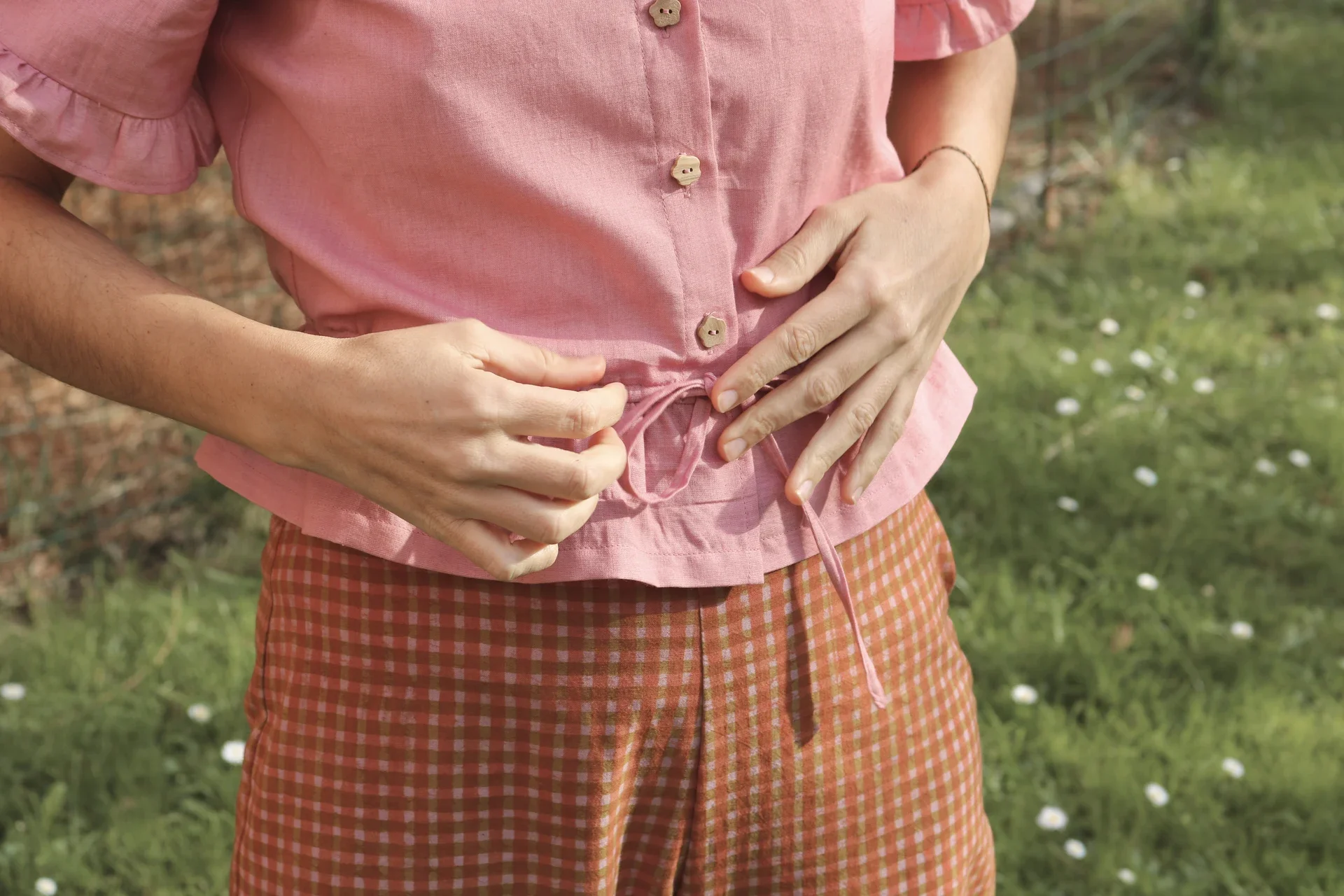 Blusa Aura de algodón orgánico en rosa de Madder Root, destacando sus botones de madera en forma de flor y el lazo ajustable en la cintura.