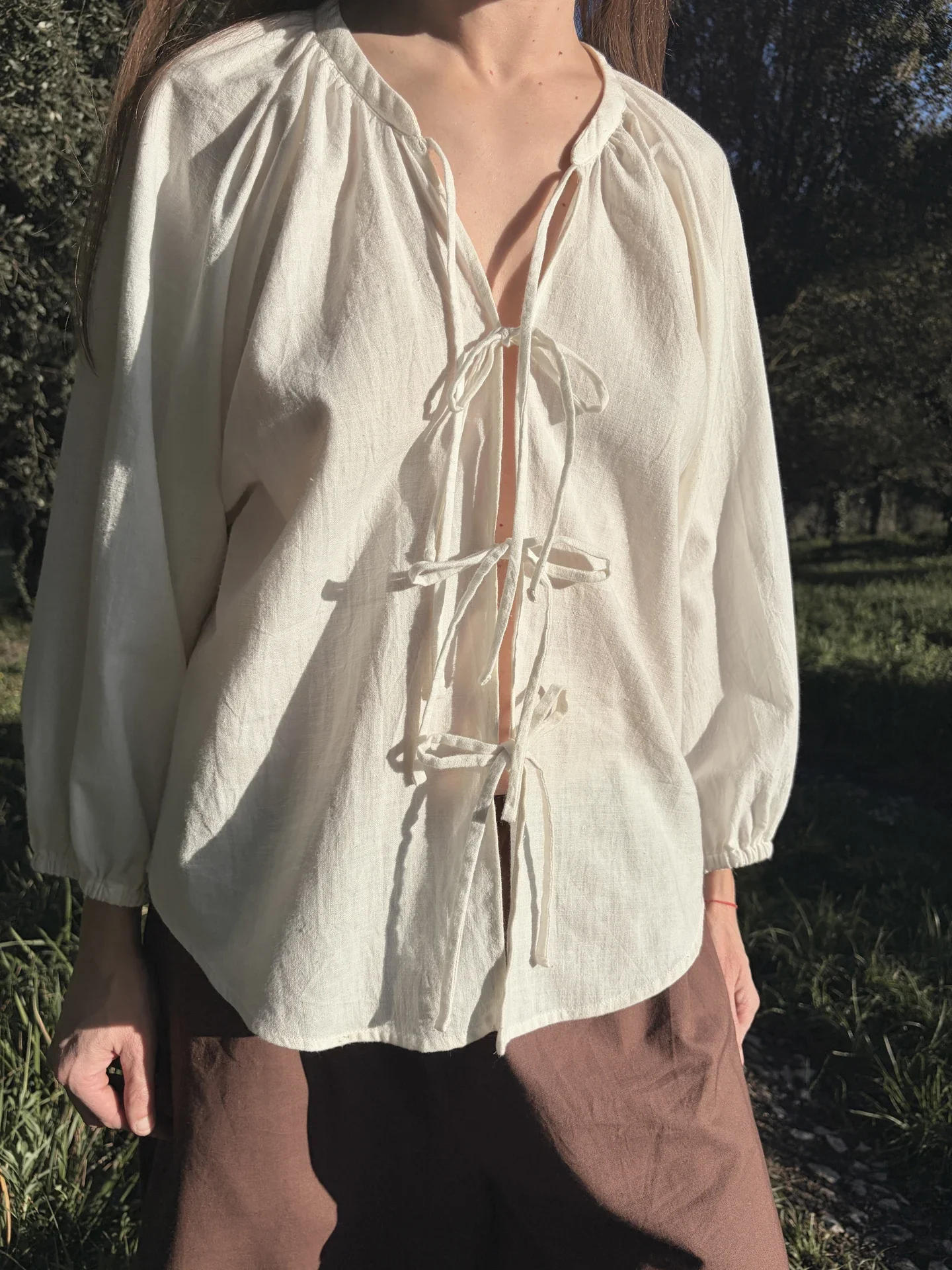 Mujer vistiendo la Blusa Cala blanca de algodón orgánico con lazos delanteros, sosteniendo flores silvestres. Moda sostenible confeccionada bajo pedido.