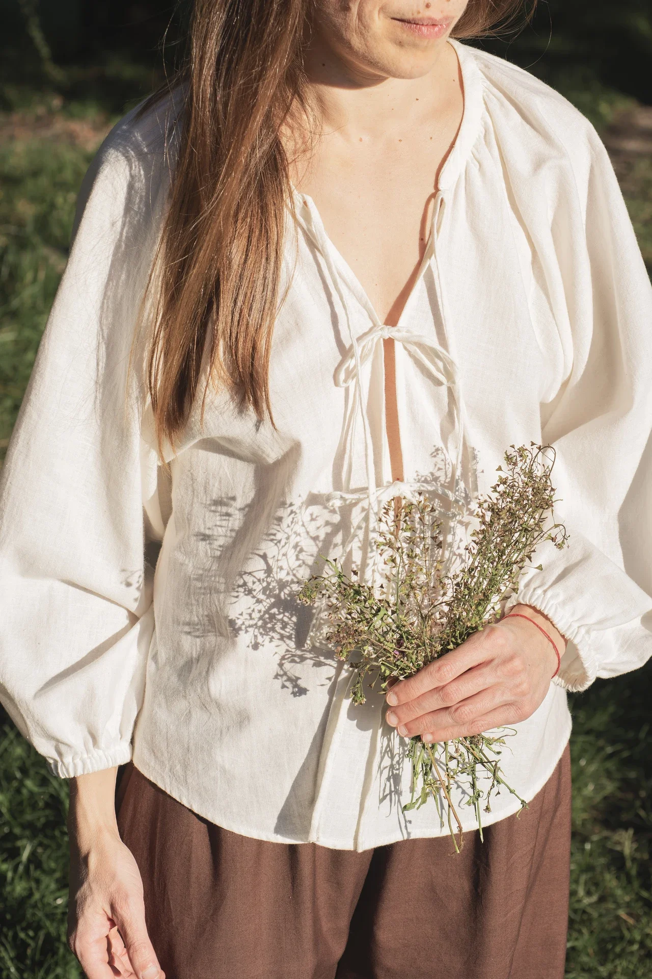Mujer vistiendo la Blusa Cala blanca de algodón orgánico con lazos delanteros, sosteniendo flores silvestres. Moda sostenible confeccionada bajo pedido.