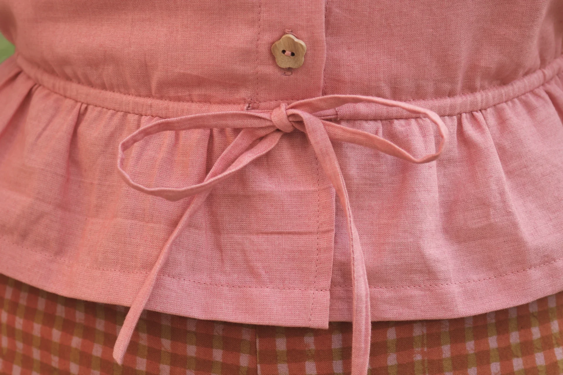 Blusa Aura de algodón orgánico en rosa de Madder Root, destacando sus botones de madera en forma de flor y el lazo ajustable en la cintura.