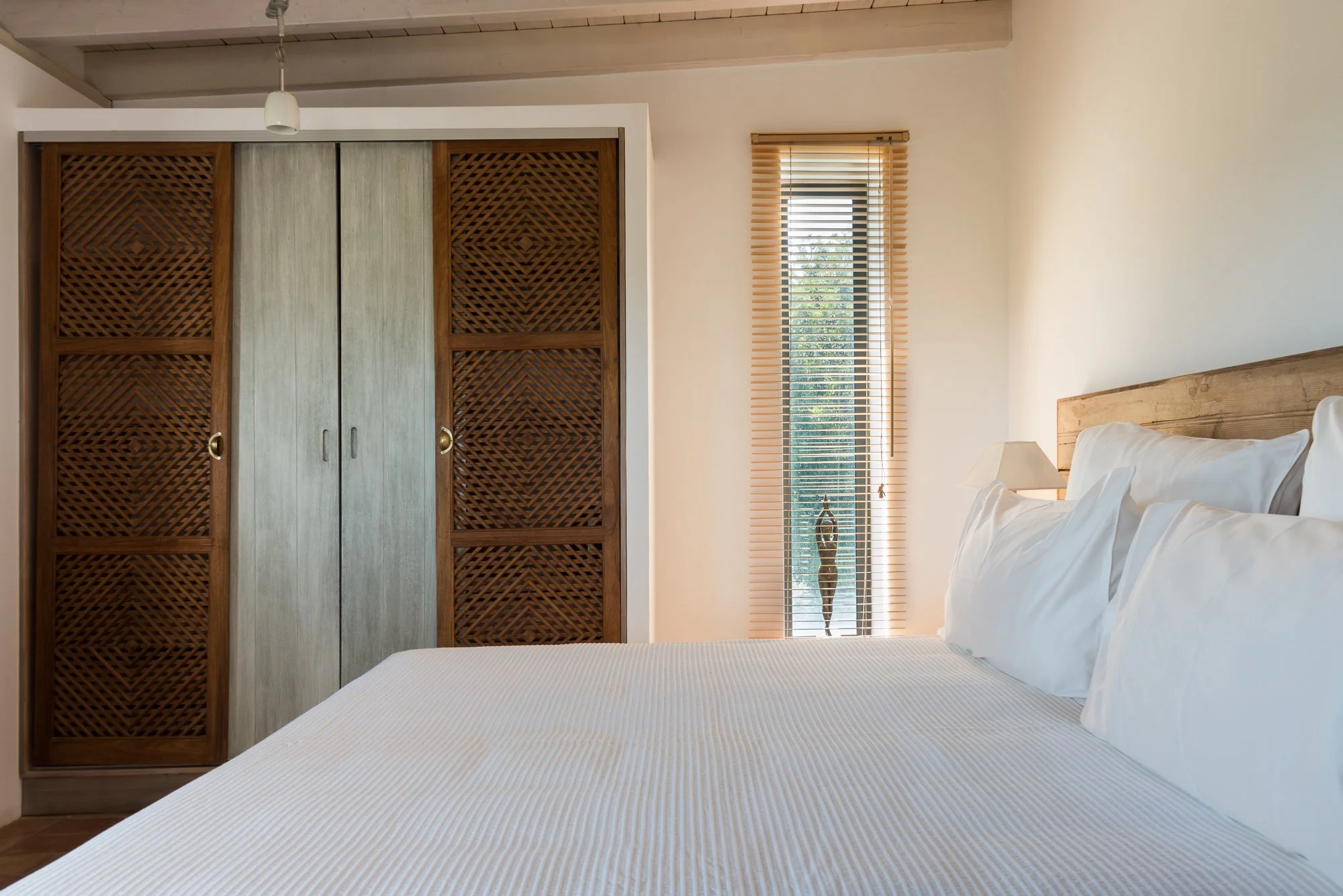 A bedroom with a bed, wooden headboard, white bedding, a next to a window with blinds, a lamp, and a wooden closet with decorative doors.