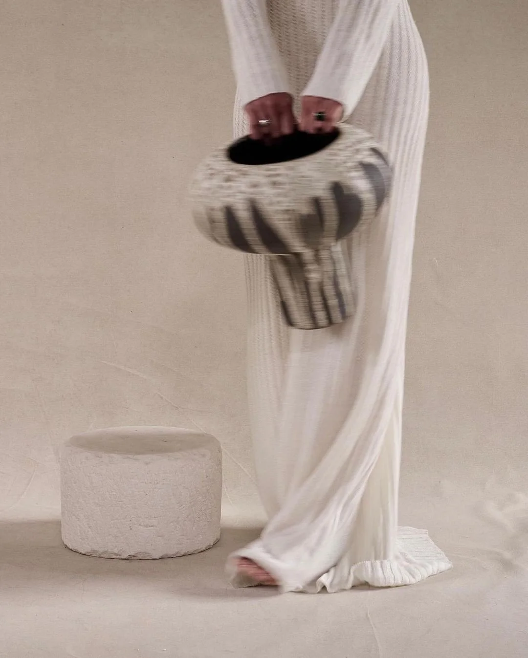 Person wearing white striped clothing holding a patterned handbag, standing near a round white textured stool against a plain beige background.
