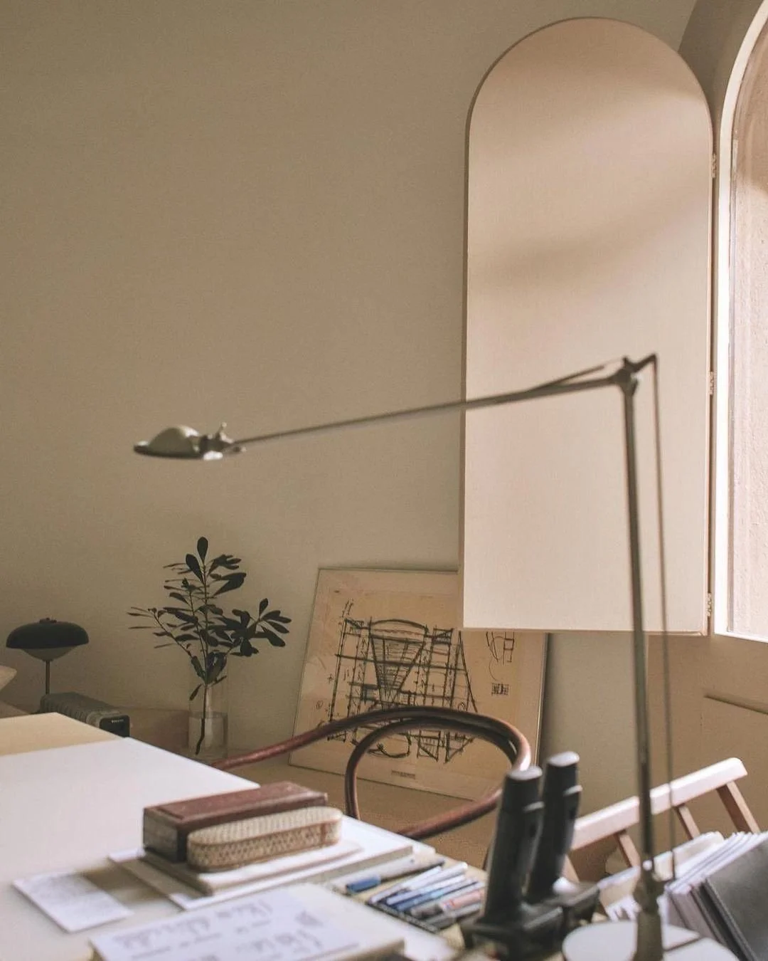 A workspace with a table, books, and a desk lamp, near a potted plant and a framed blueprint leaning against the wall. A window lets in natural light.