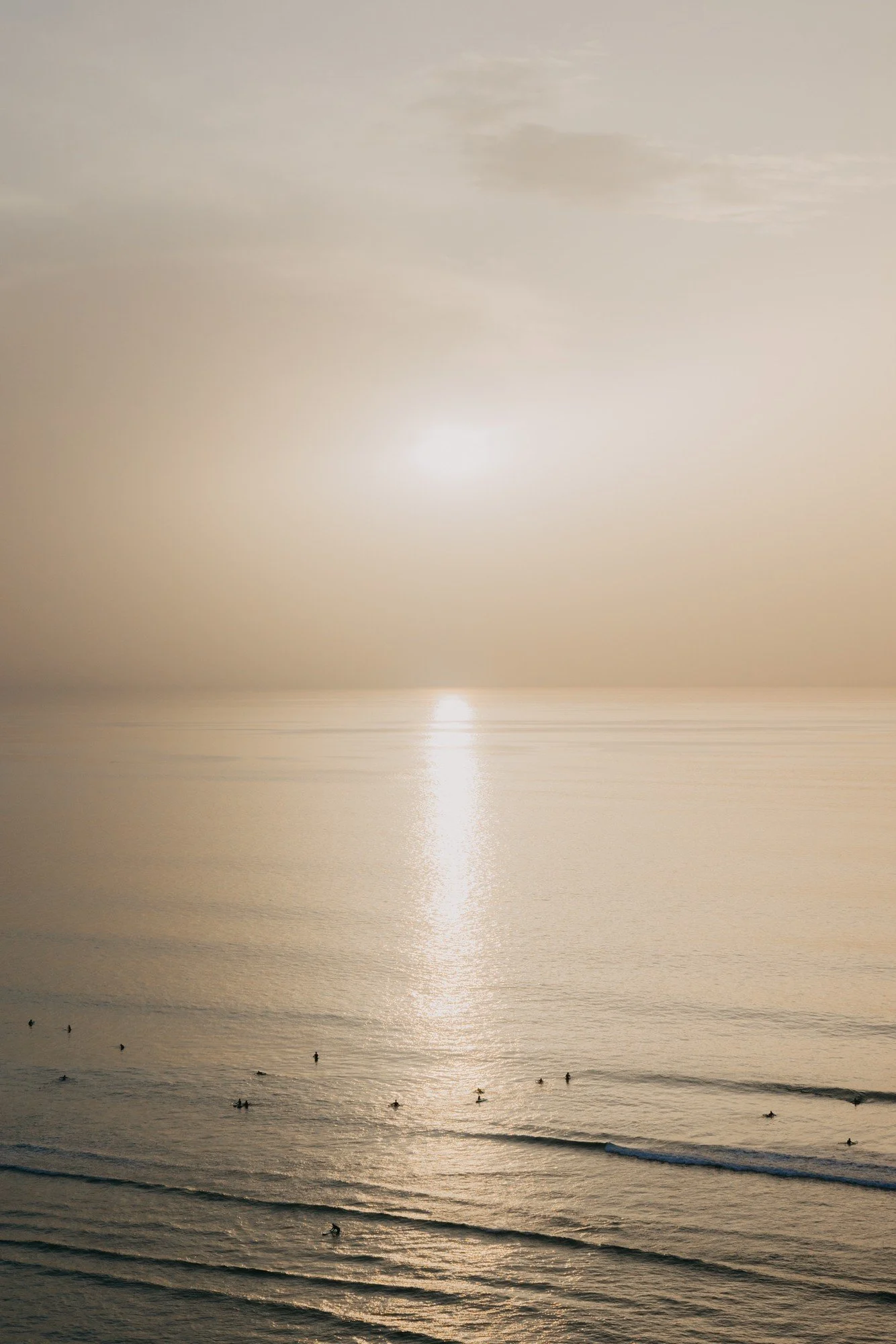 A tranquil ocean scene at sunset with the sun low on the horizon, casting a warm glow and reflecting off the water, with small waves and several surfers in the distance.