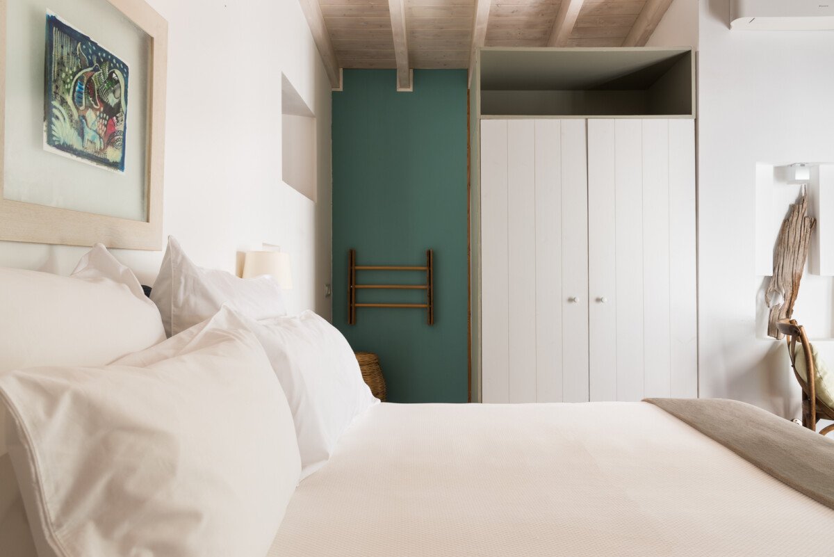 A cozy bedroom with a white bed, pillows, and a beige blanket, featuring artistic decor including a framed painting of a bird on the wall, a small wooden ladder on a teal accent wall, a white wardrobe, and a wooden chair with a draped piece of fabric.