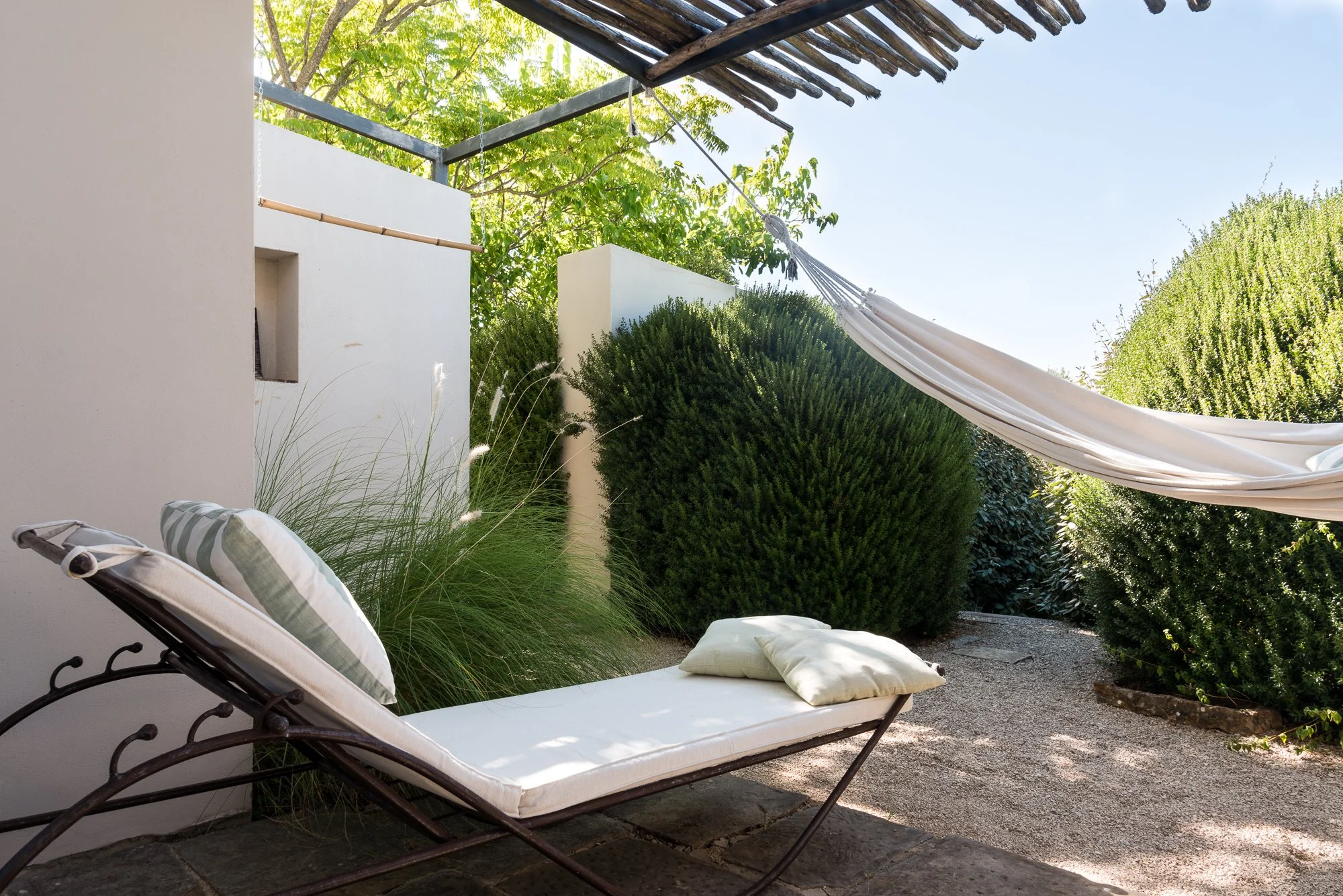 Outdoor patio with a white cushioned lounge chair with a pillow, tall grass plants, green bushes, and a white hammock under a wooden pergola.