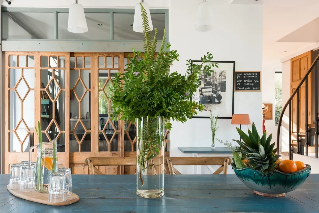 Indoor dining area with a wooden table, a large vase of green leafy plants, a bowl of fruit, and a tray with glasses and a pitcher of water with lemon.