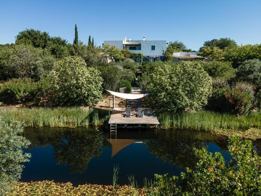 A lakeside yard with graffiti on a modern white house in the background, surrounded by lush green trees and bushes, and featuring a dock with lounge chairs and a deck chair, reflected in the calm water.