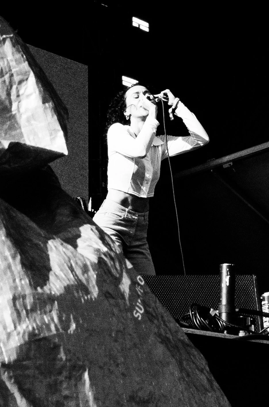 A woman singing into a microphone on stage during a performance, with stage equipment visible in the foreground in black and white.
