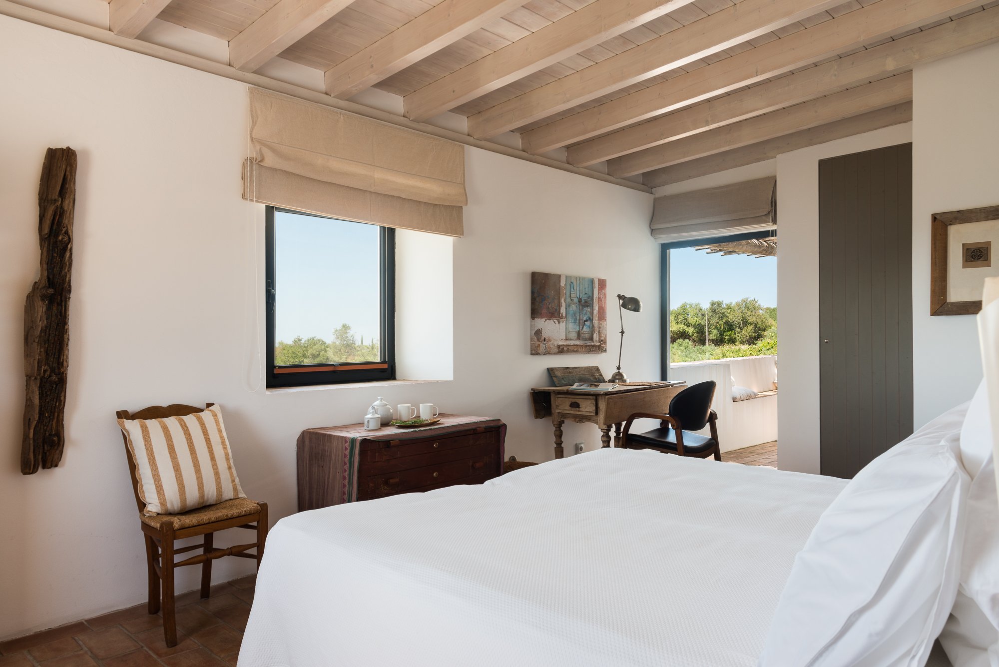 A cozy bedroom featuring a white bed, wooden ceiling beams, and windows with beige blinds overlooking greenery, with an antique wooden dresser, a black desk chair, and a small table on a balcony.