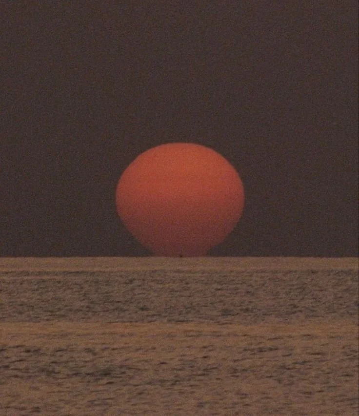 A large red sun setting over the ocean with dark clouds in the background.