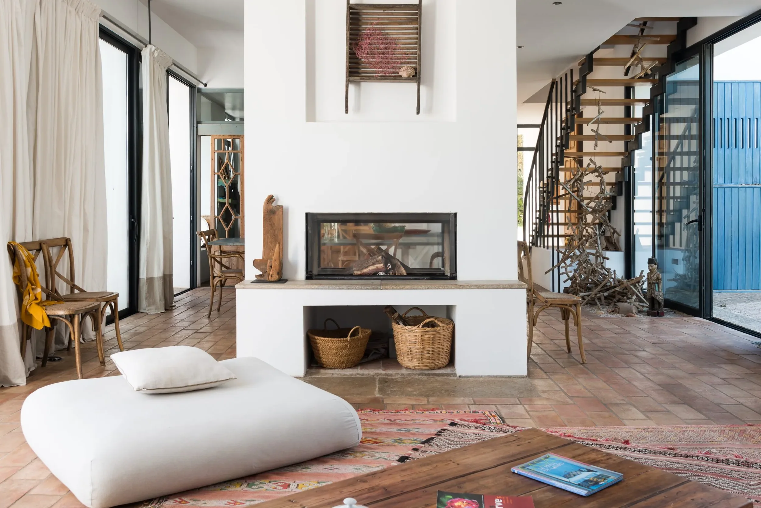 Modern living room with a white fireplace, wicker baskets, and wooden furniture, featuring sliding glass doors and a staircase with wood and black metal railing.