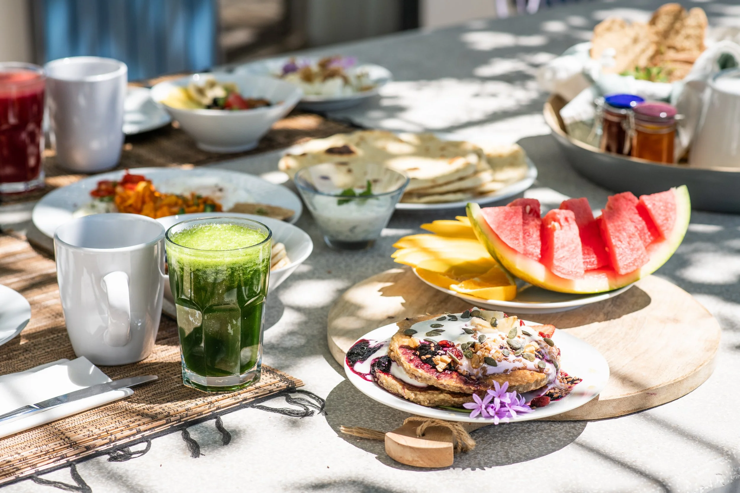 A table set for breakfast with plates of watermelon slices, pancakes with syrup and toppings, a green drink, a white mug, bowls of fruit and other breakfast foods, and cups of tea or coffee, with sunlight and shadows.