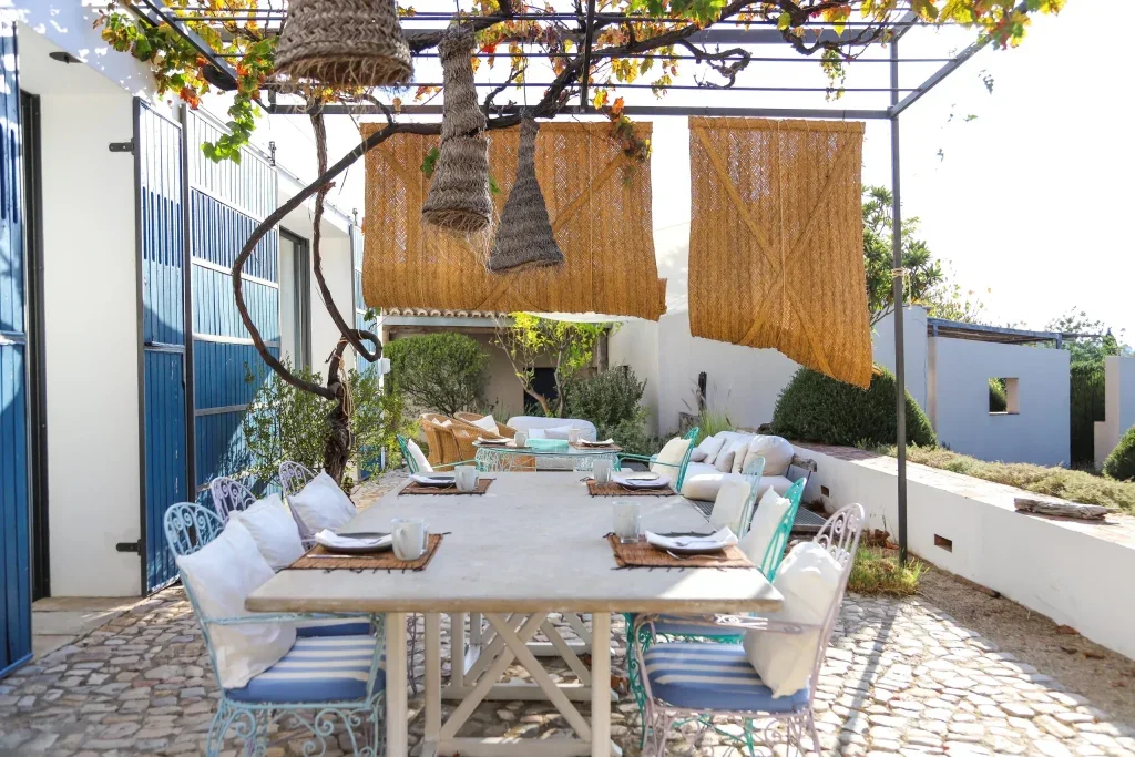 Outdoor dining area with a long table set with plates, cups, and cutlery, surrounded by white and turquoise chairs. Overhead is a pergola with hanging woven decorations, shaded by bamboo panels, in a sunny garden setting with cobblestone pavement and green bushes.