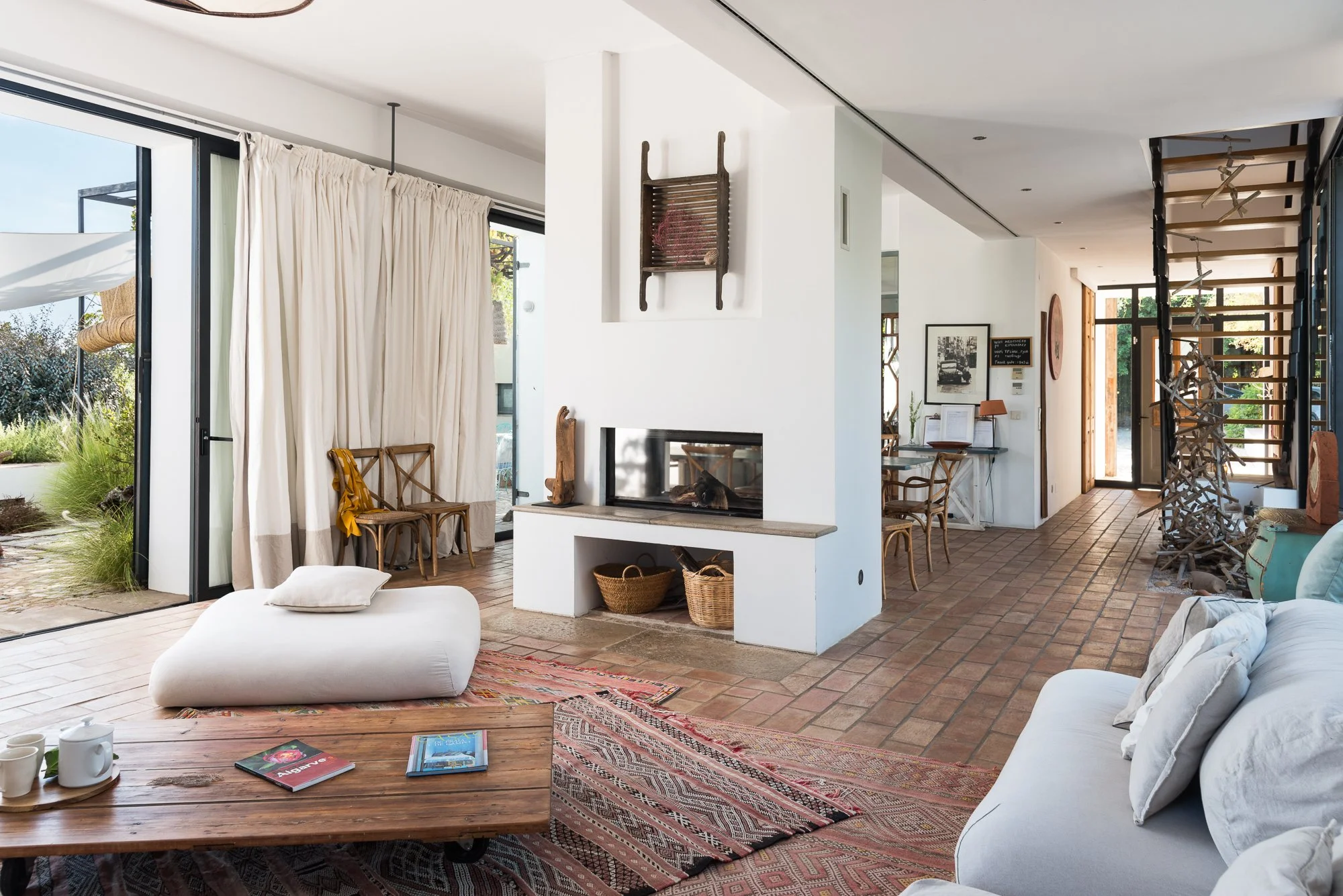 Living room with white walls and brick flooring, featuring a fireplace, a white sofa, a wooden coffee table with books and cups, a patterned rug, and large windows with curtains showing a garden outside.