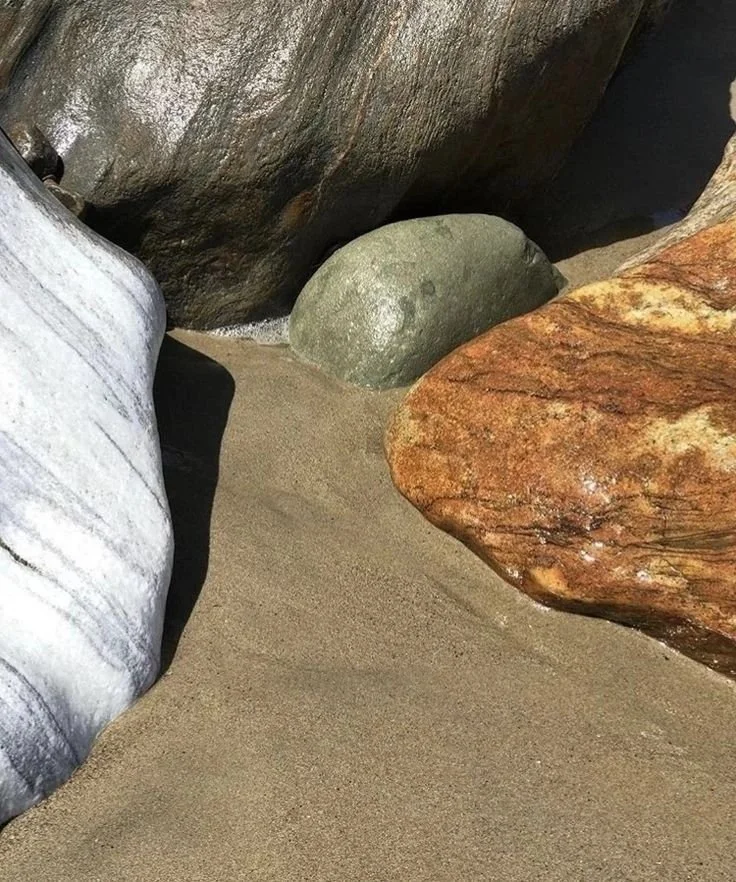 Close-up of a sandy area with four large rocks of different colors and textures, with rocks and shadows nearby.