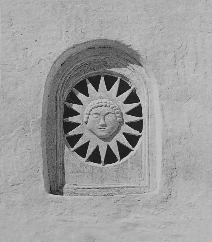 A stone wall with an arched window containing a decorative sun motif with a human face at the center.
