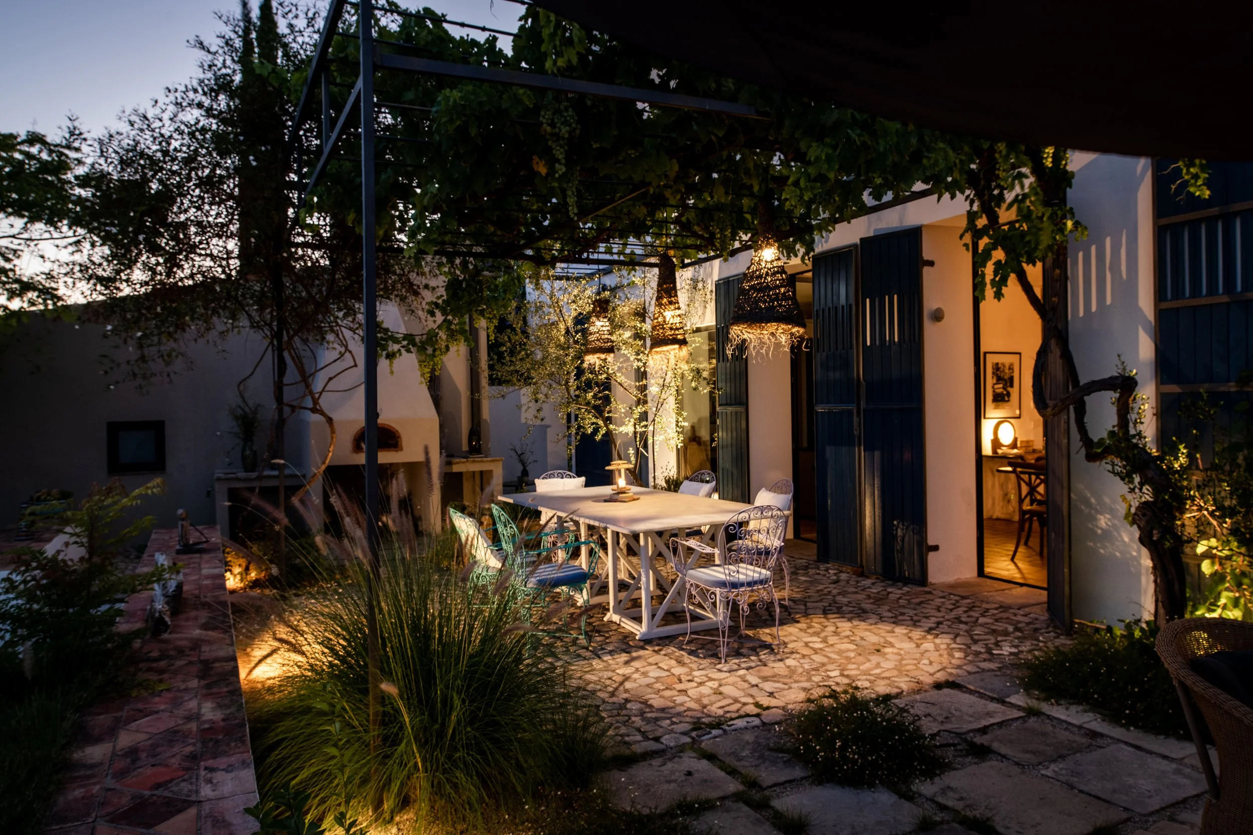 Outdoor dining area with a long white table, white and blue chairs, warm lighting, and a vine-covered pergola at dusk.
