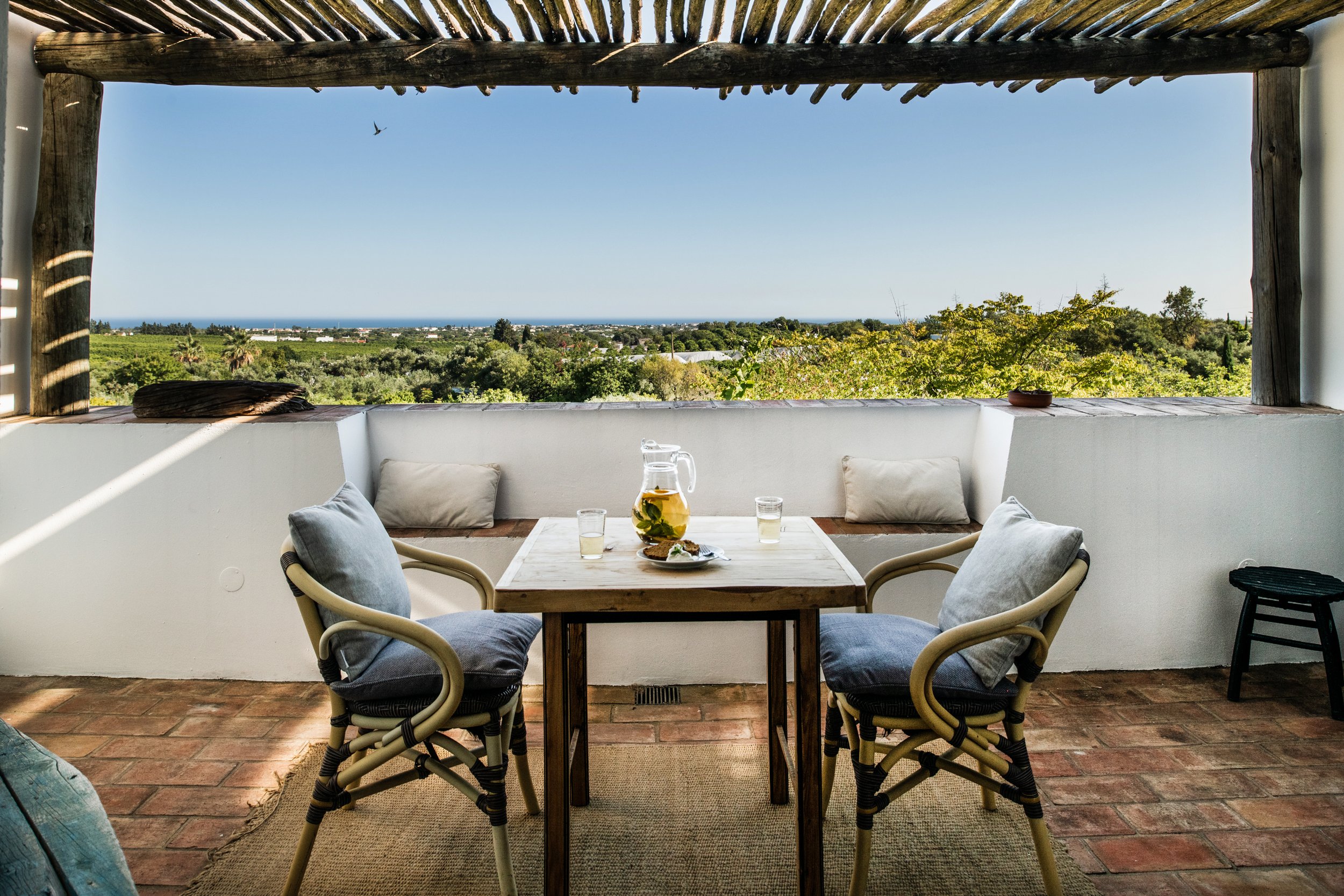 Outdoor patio with a wooden table set for drinks, surrounded by chairs with cushions, overlooking a lush green landscape and distant ocean under a blue sky.