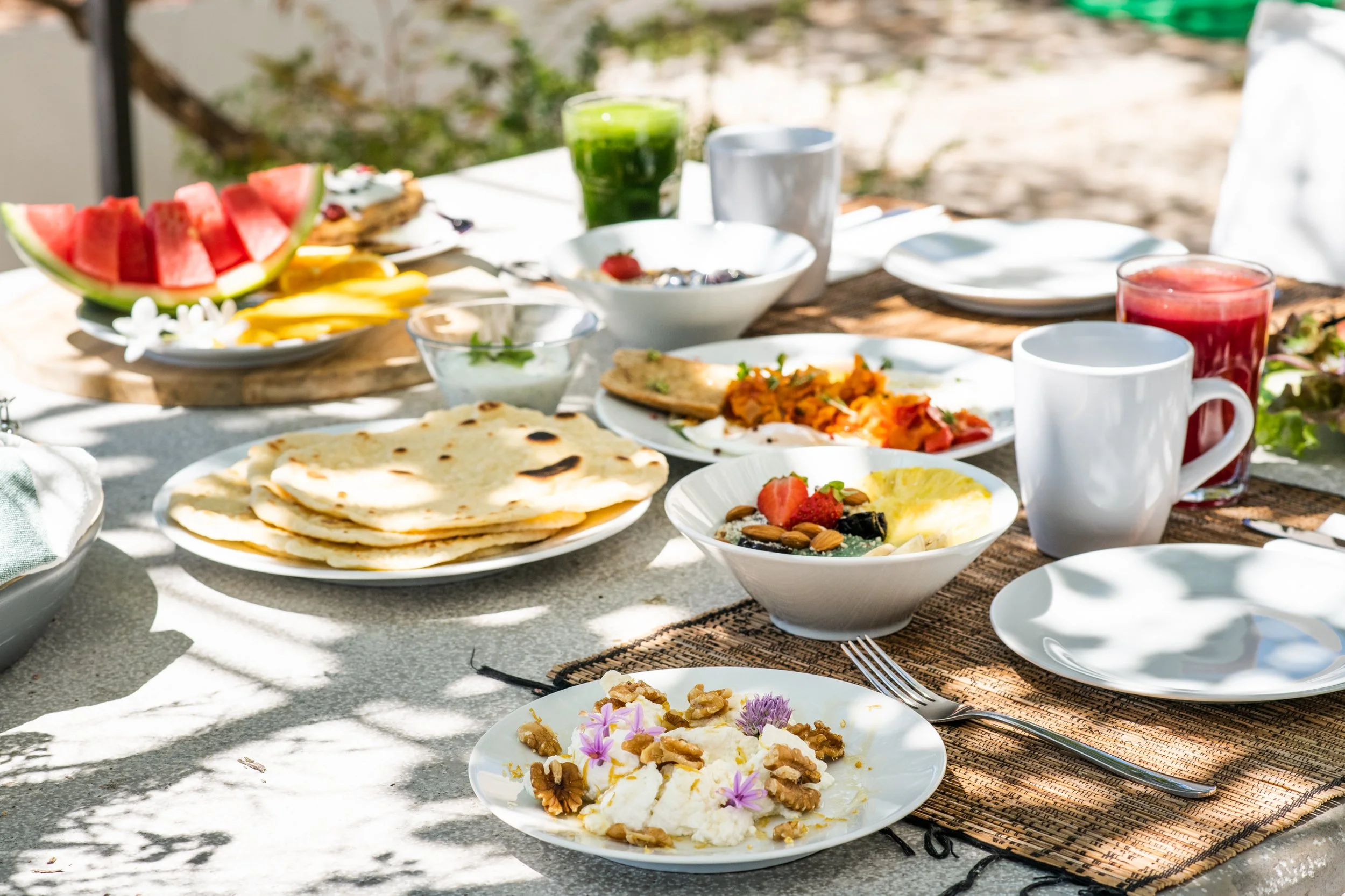 A table set for breakfast with various dishes including watermelon, mango, flatbread, yogurt with nuts and flowers, fruit salad, and a colorful vegetable dish, accompanied by glasses of juice and coffee mugs, on a outdoor patio surrounded by greenery