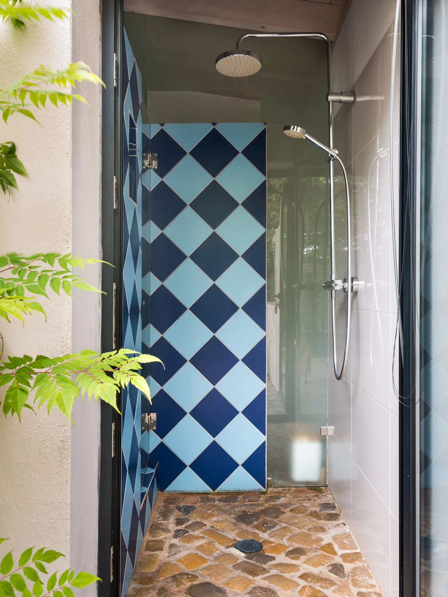 An outdoor shower with two showerheads, one fixed and one handheld, mounted on a wall. The back wall has blue and light blue diamond-patterned tiles. The floor is made of irregularly shaped stones. A glass door or partition separates the shower area 