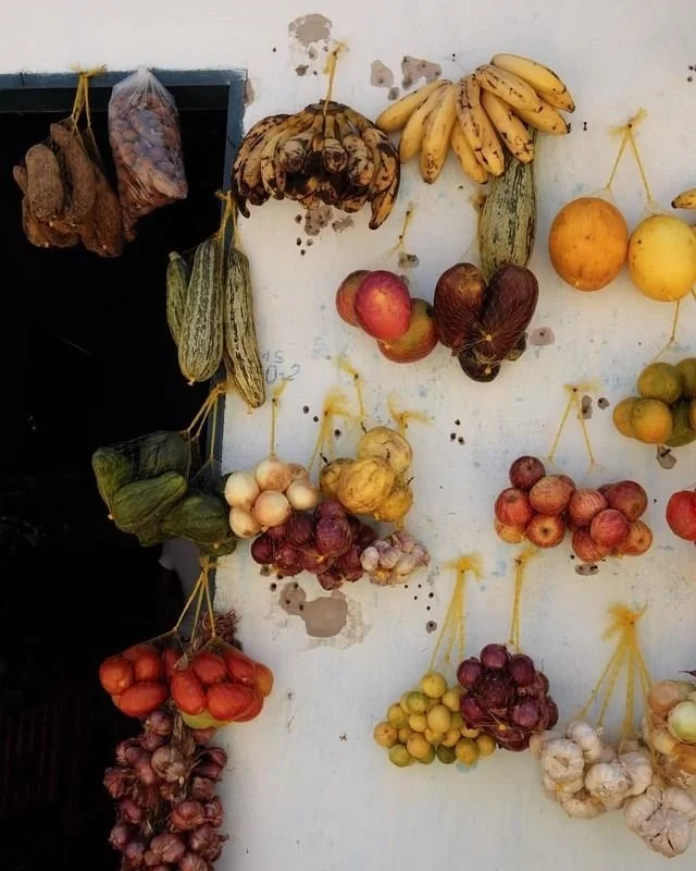 Various fruits including bananas, apples, onions, garlic, and other produce hanging on a white wall, with some in plastic bags.