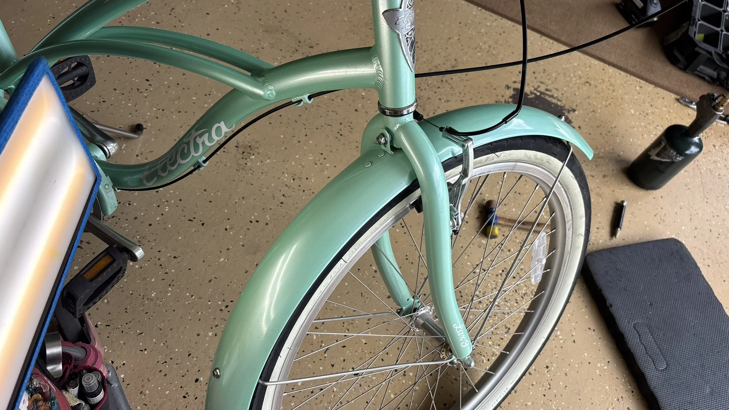 Close-up of a mint green cruiser bicycle with a front fender, pedal, and part of the frame visible, sitting on a tan speckled floor in a workshop or garage.