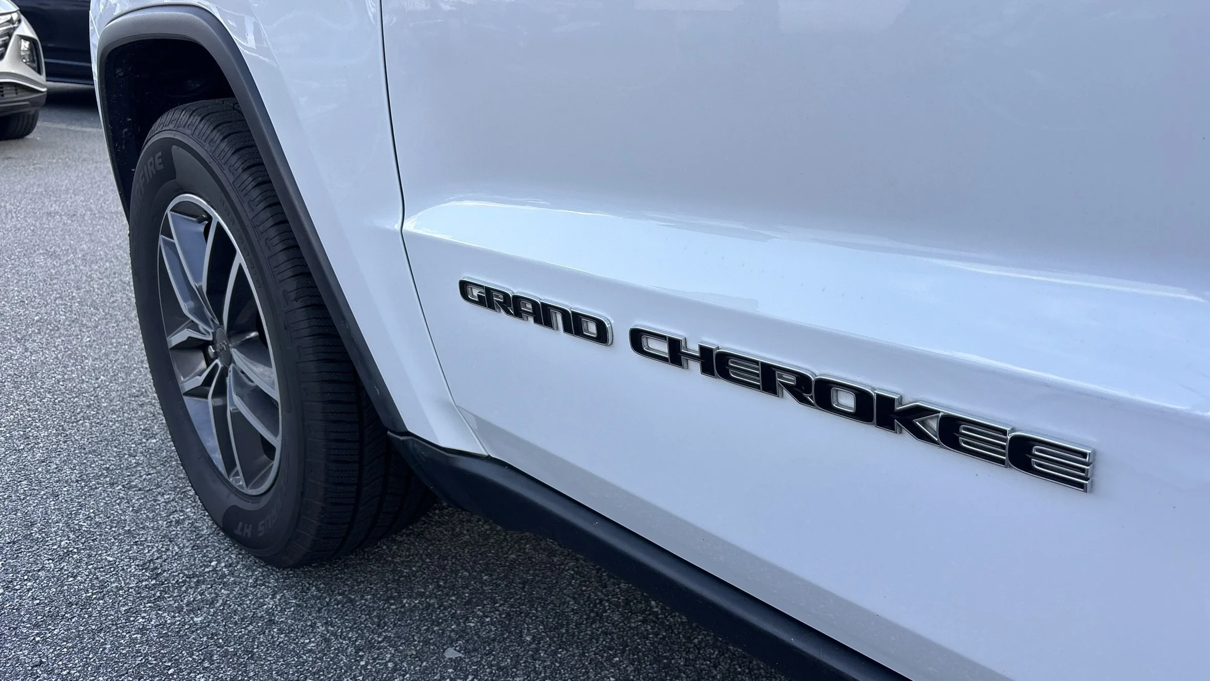 Close-up of a white vehicle's side with the badge 'Grand Cherokee'. The front wheel and part of the tire are visible, parked on a textured surface.