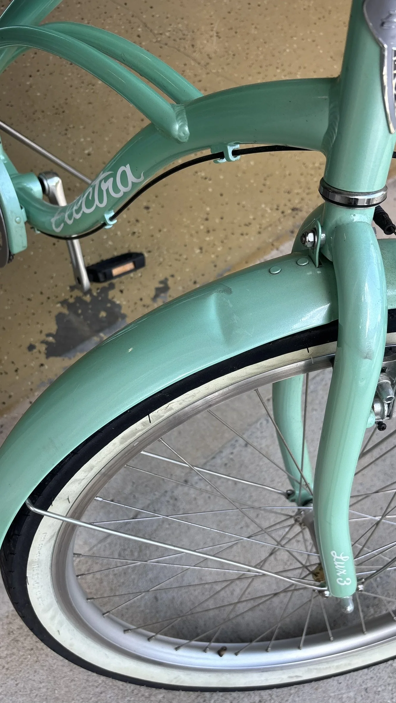 Close-up of a mint green Penny-farthing style bicycle, showing the front wheel, fender, and part of the frame with the words "Pacha" and "Luv 3" written on it.