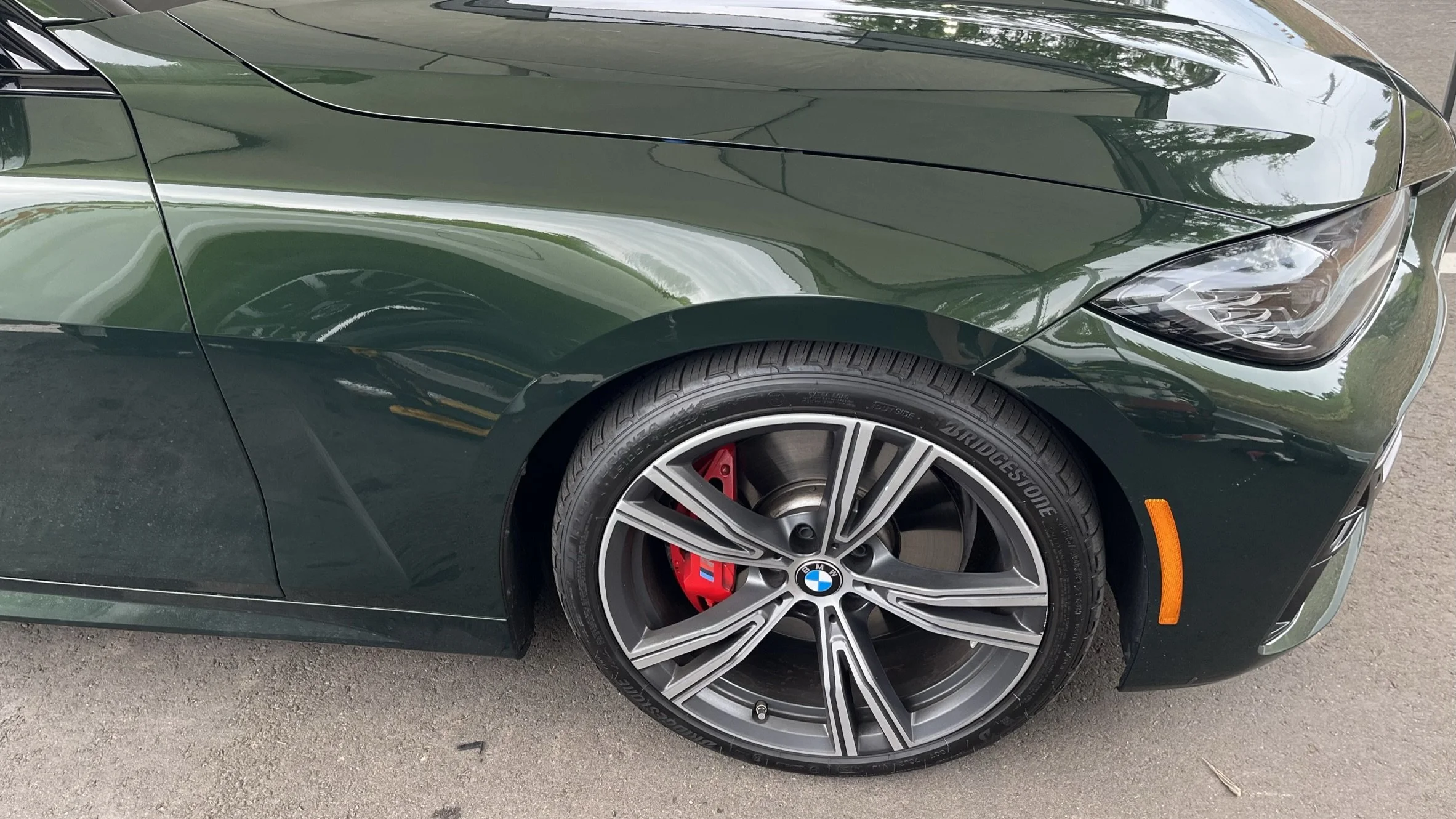 Close-up of the front part of a dark green BMW sports car showing the front wheel, tire, red brake caliper, and sleek headlight.