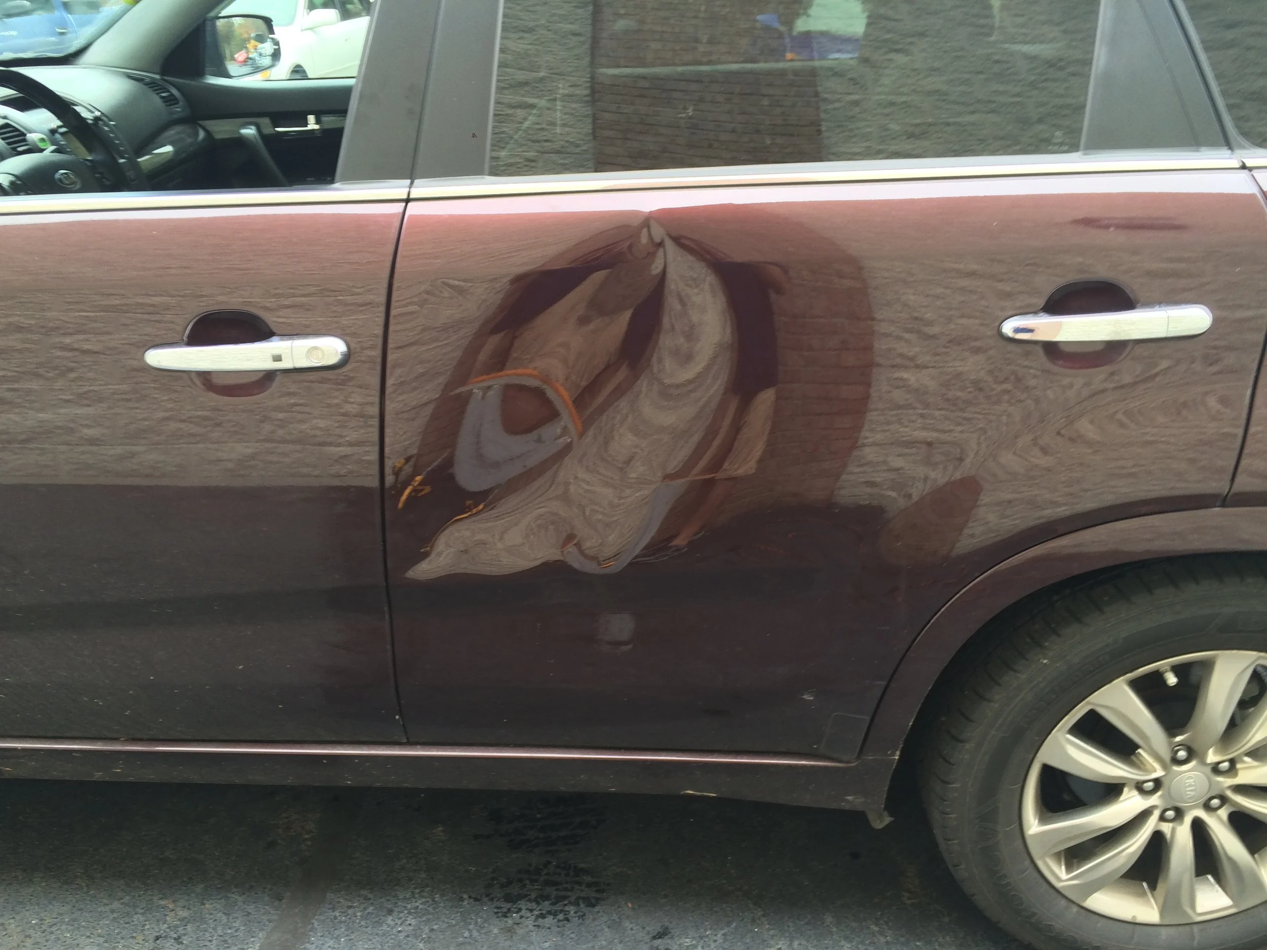 Car door with noticeable dent and scratches on the driver's side, reflecting the surrounding area.