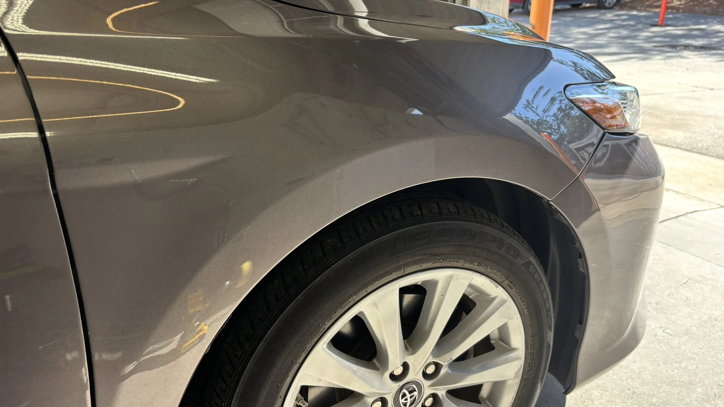 Close-up of the front left side of a gray Toyota sedan, showing the wheel, tire, front fender, headlight, and a dent on the fender.