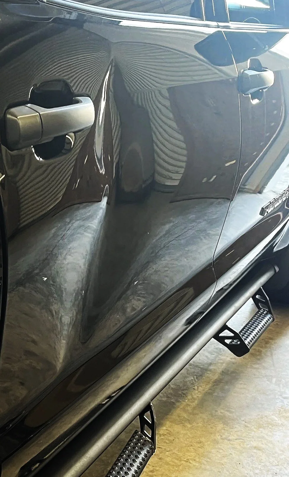 Close-up of a black car's side door with a metallic handle and reflection of a person wearing a striped shirt, alongside a black running board.