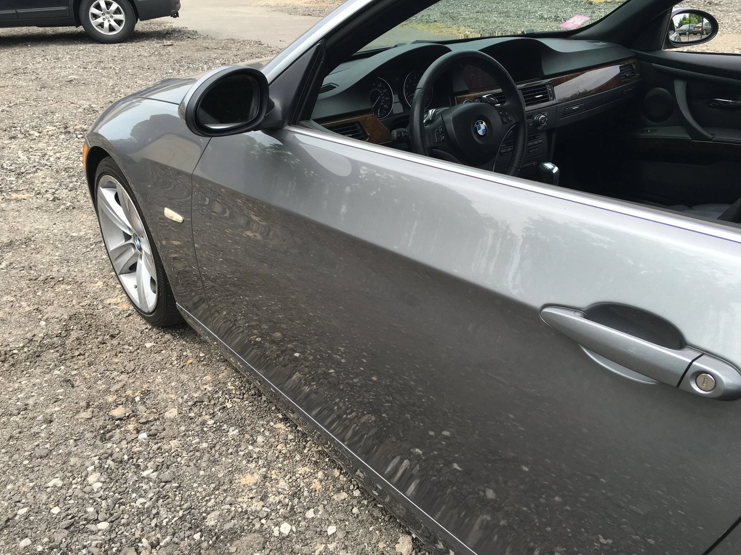 Close-up of a gray BMW coupe parked on gravel, showing the front left side including the wheel, side mirror, and interior dashboard through the window.