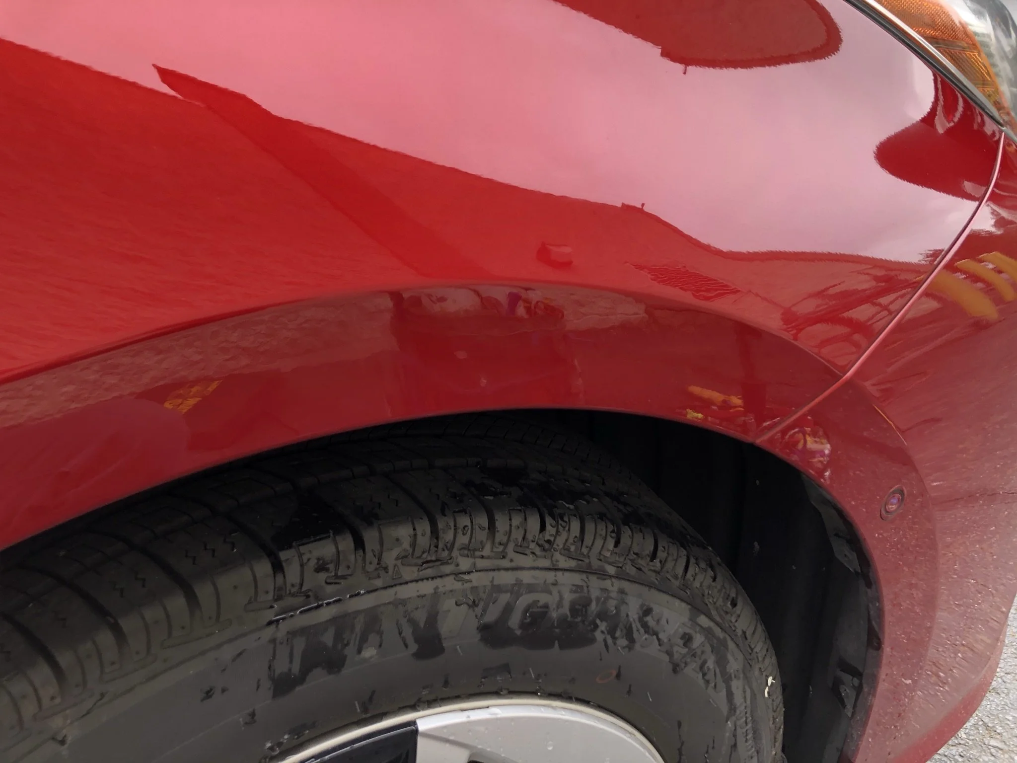 Close-up of a red car's front wheel and fender, showing the tire with tread pattern and the shiny red paint, with reflections of the surroundings.