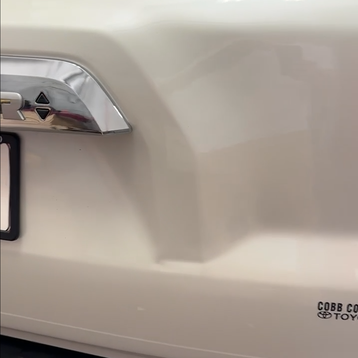 Close-up of a metallic car badge with a black triangle symbol on a cream-colored vehicle, partially showing the license plate and the logo of Cobb County Toyota.
