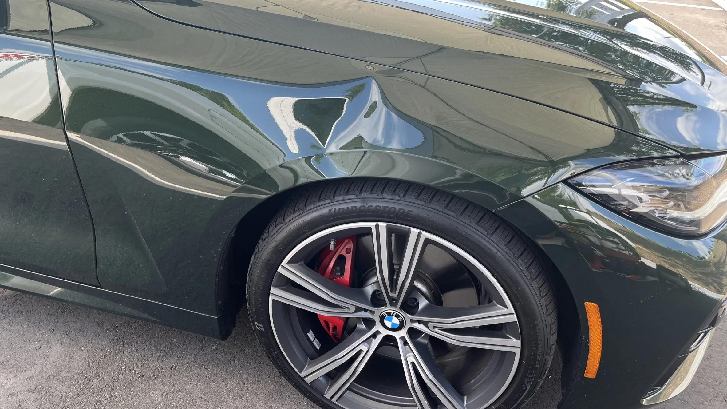 Close-up of a dark green BMW car with a significant dent on the front left fender above the wheel, which has a red brake caliper and silver alloy rim, parked on a paved surface.