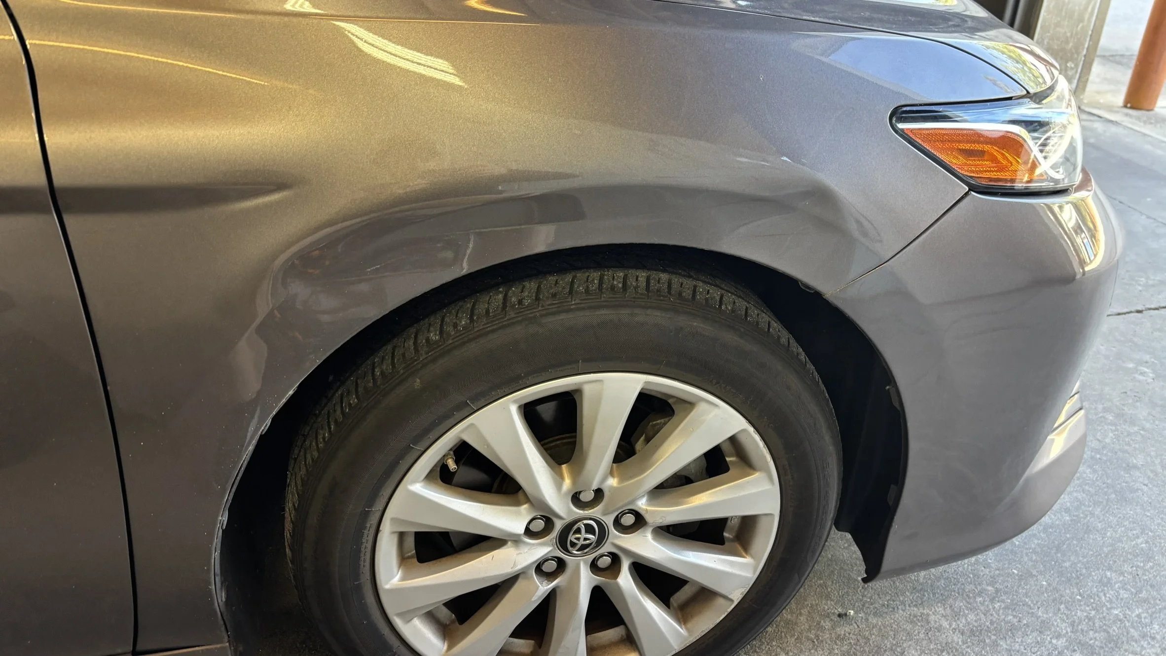 Close-up of the front left side of a gray Toyota sedan, showing the wheel, tire, and part of the headlight and fender.