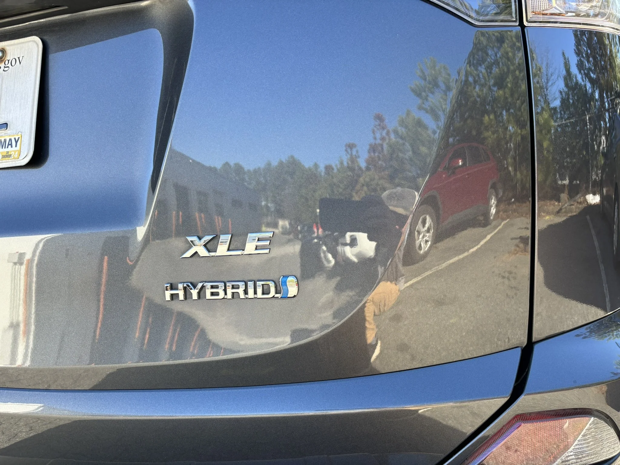 Close-up of the rear of a black hybrid vehicle showing the badges 'XLE' and 'HYBRID' on the trunk, with reflections of the surroundings and other cars visible in the glossy paint.