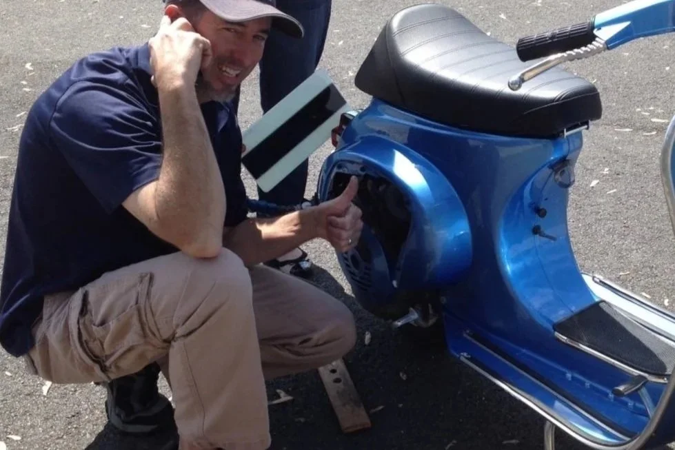 A man crouching next to a blue scooter, giving a thumbs up while talking on a cell phone.