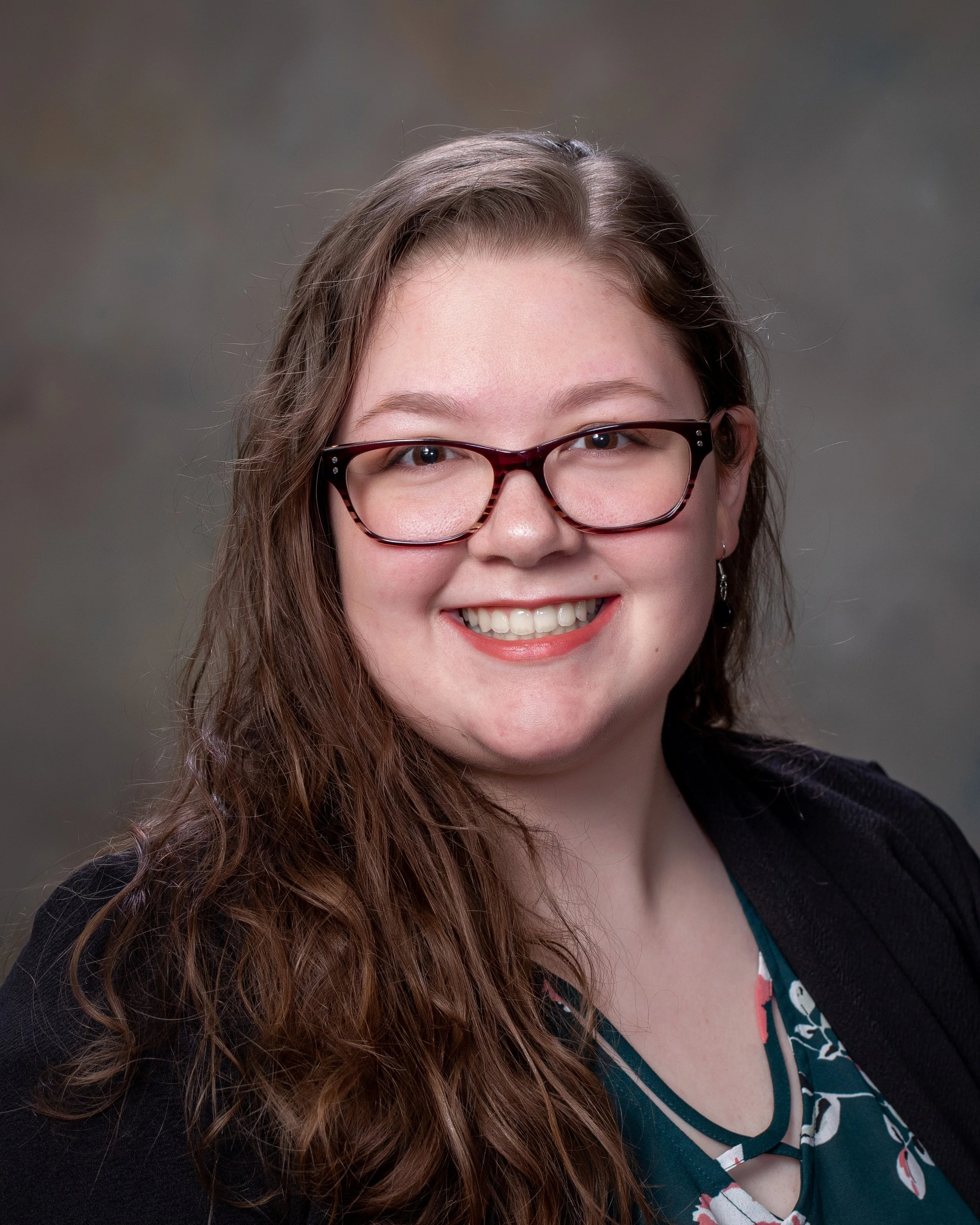 Portrait of a young woman with long curly hair, glasses, and a smile, wearing a black blazer and a patterned blouse, against a neutral background.