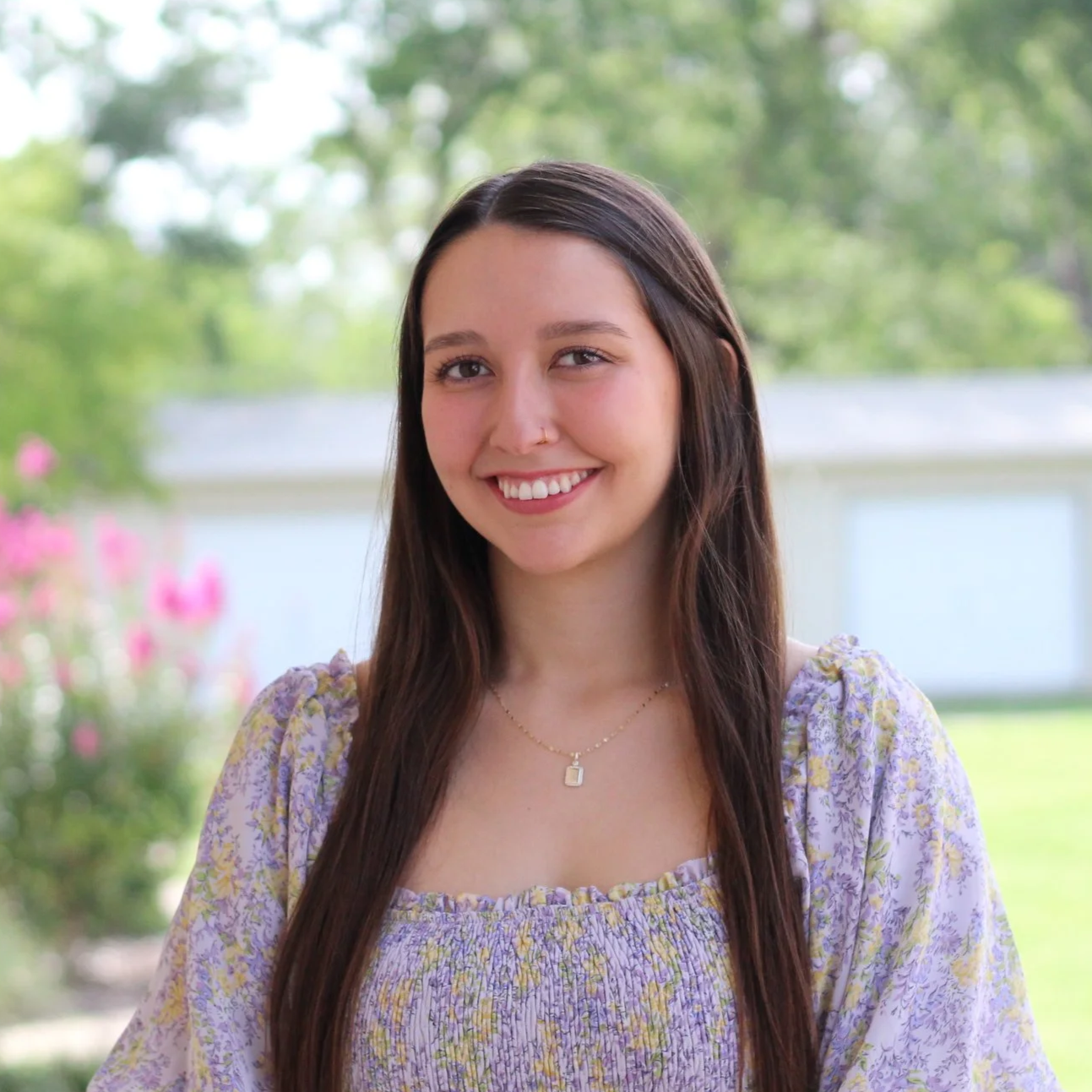 Young woman outdoors with long brown hair, smile, wearing a purple floral dress and a necklace, background of green trees and a white fence.