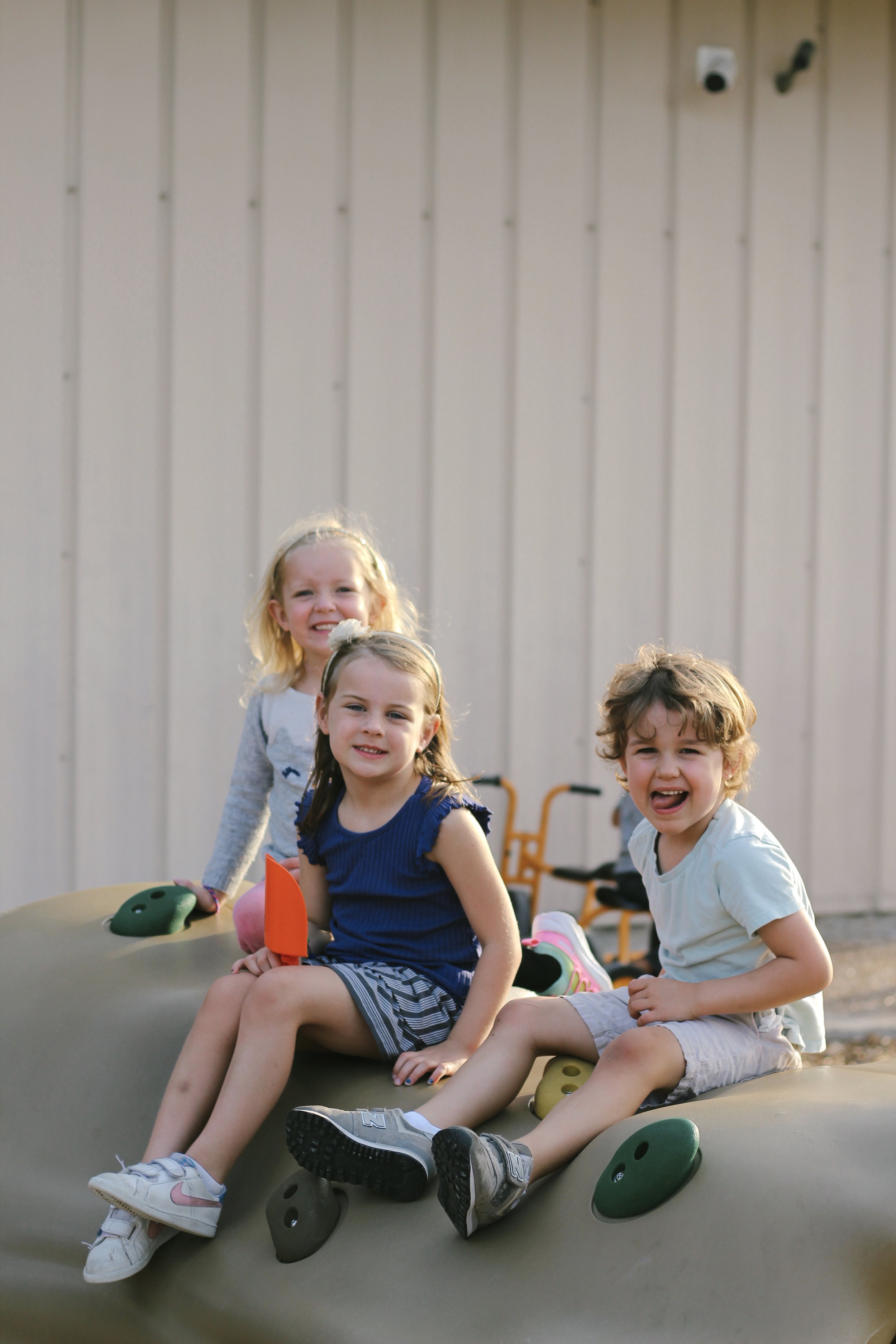 Three children sitting on a large climbing rock, smiling and playing outdoors.