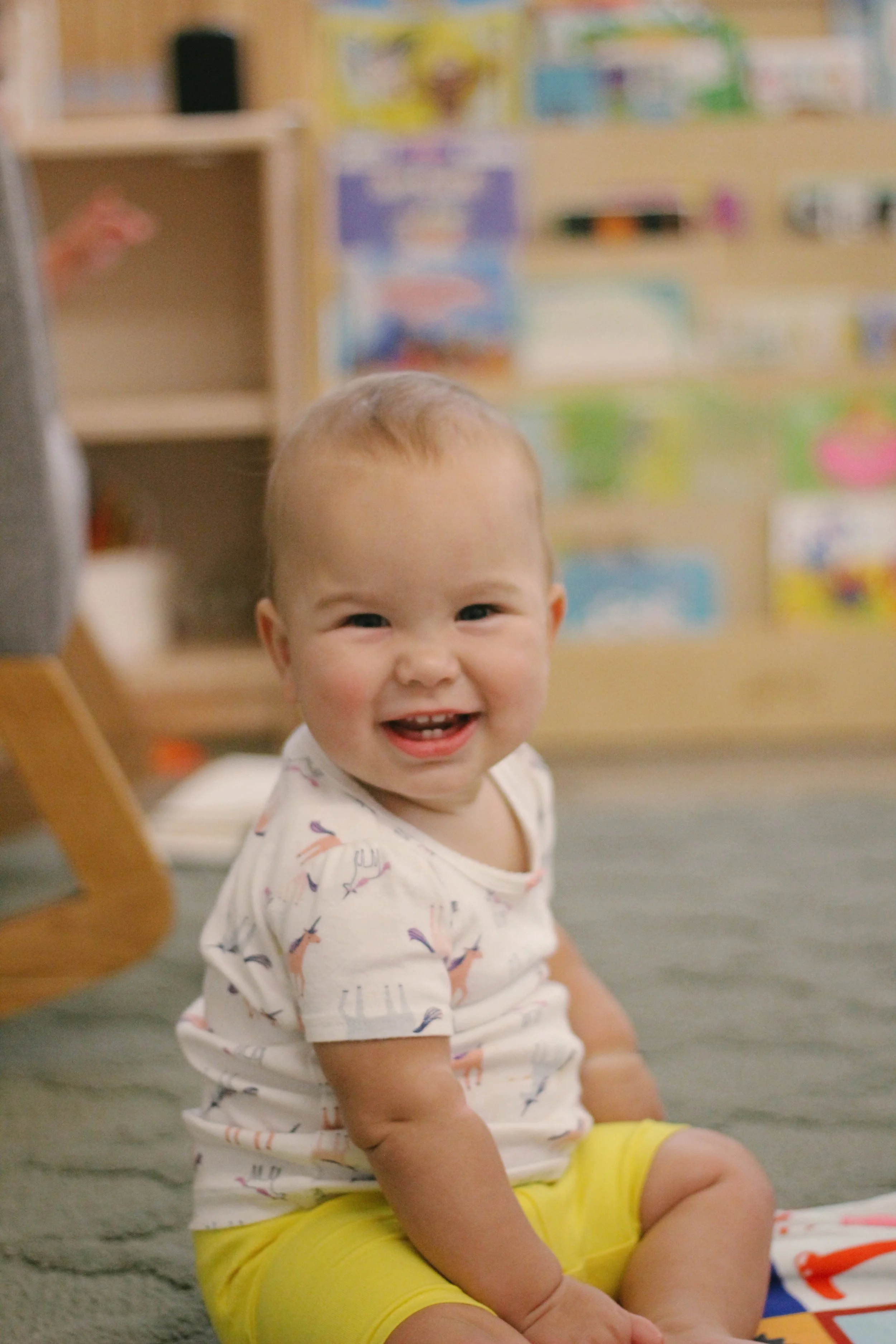 An infant sitting on the floor in a colorful room, playing with other infants.