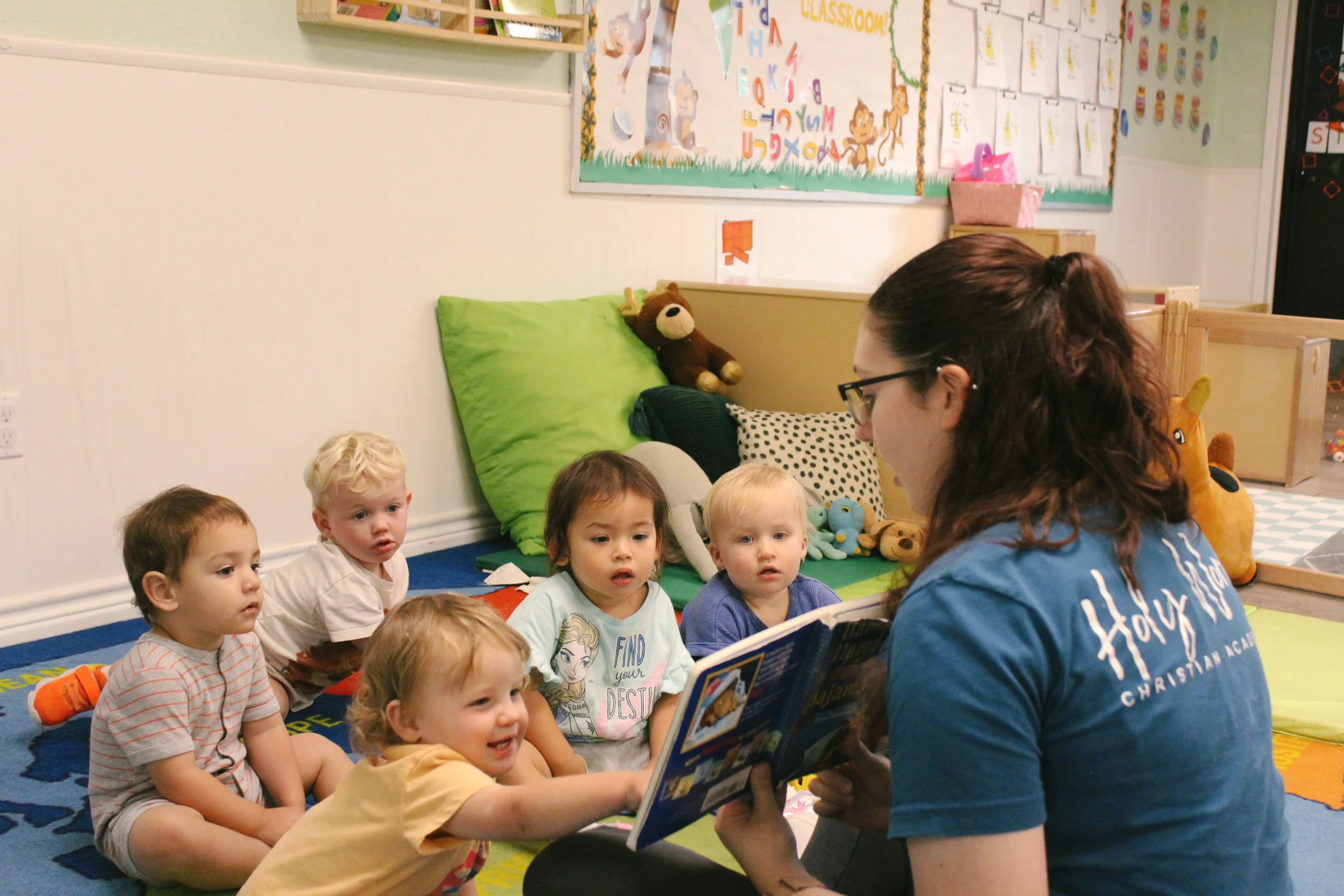 A group of five young children sitting on a colorful carpet in a classroom, listening to a teacher reading a book. The classroom has plush pillows and stuffed animals, and a wall decorated with learning posters and charts.