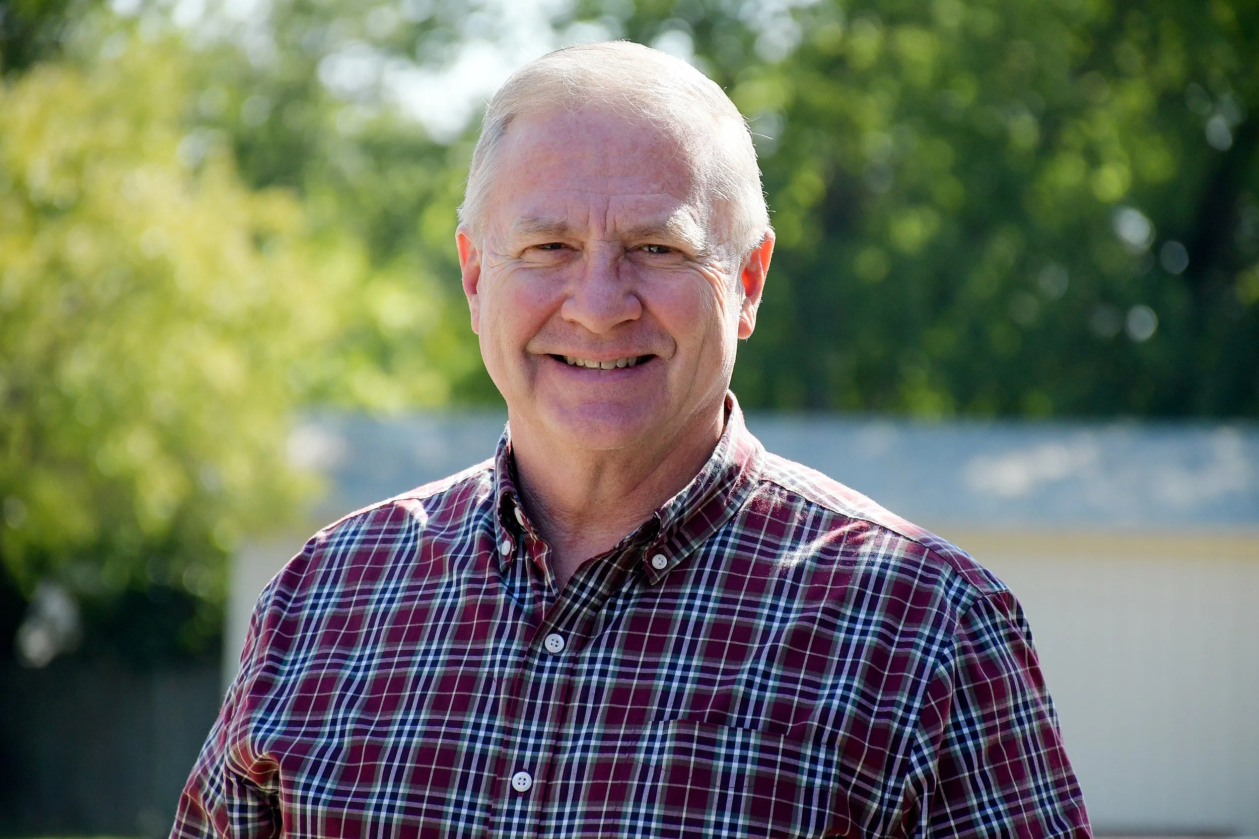 Smiling elderly man with gray hair outdoors on a sunny day, wearing a plaid button-up shirt, with green trees in the background.