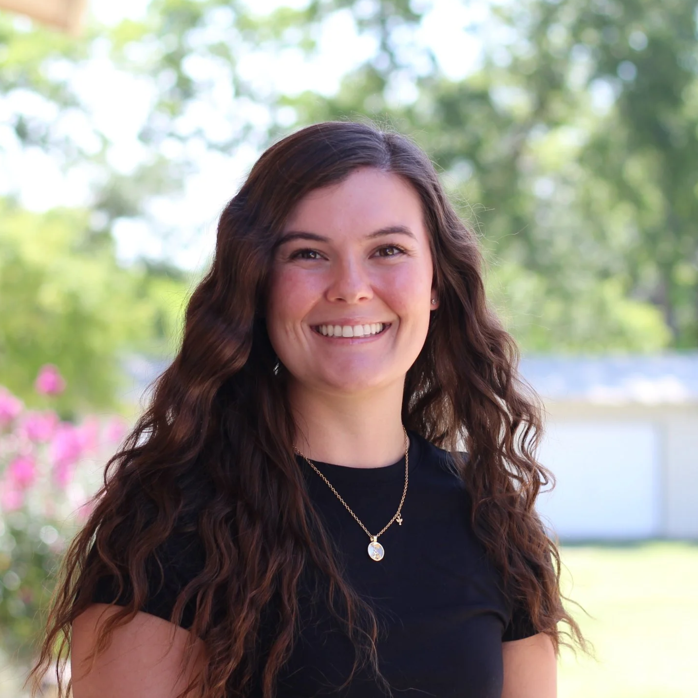 Young woman with long, curly brown hair smiling outdoors in front of trees and a white fence, wearing a black top and gold necklace.