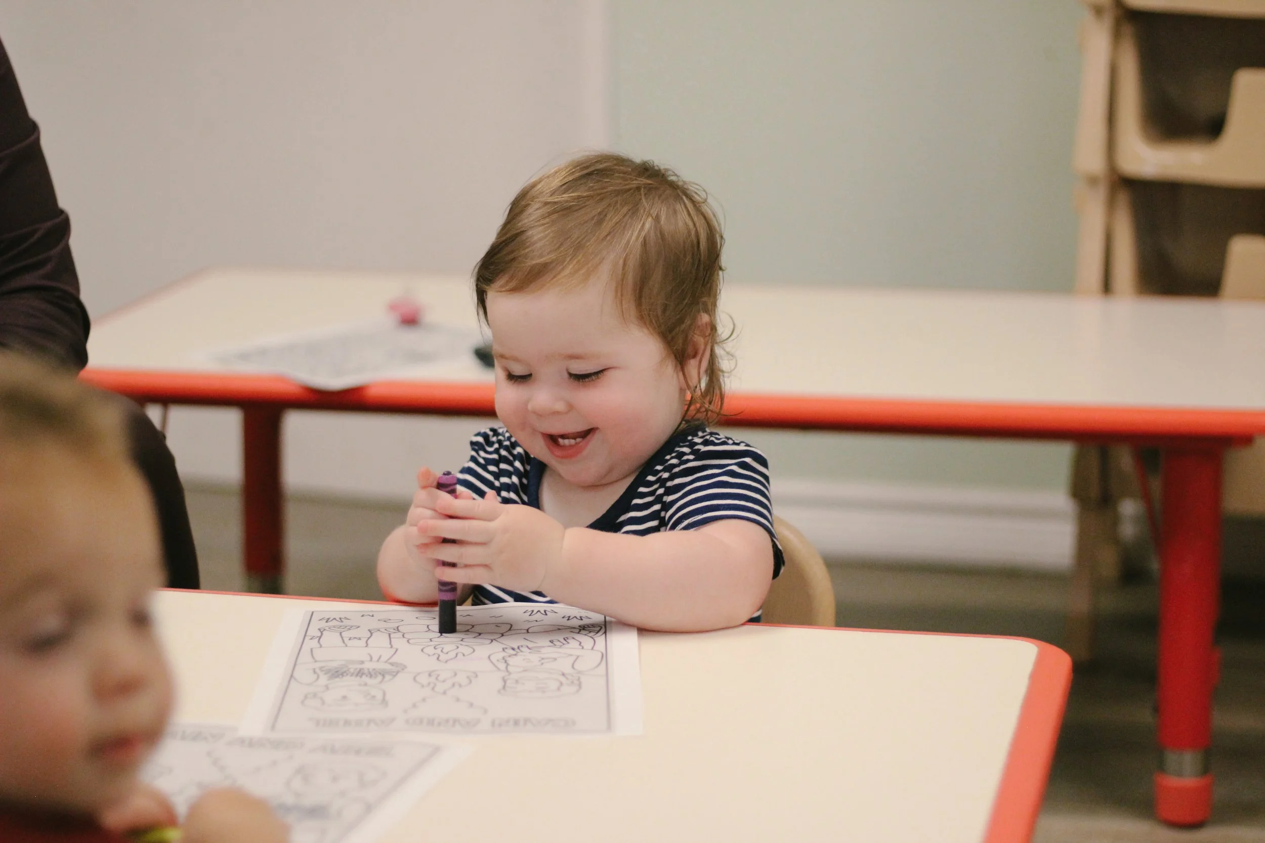 A young child coloring on a sheet of paper with a black crayon, smiling and sitting at a table in a classroom.
