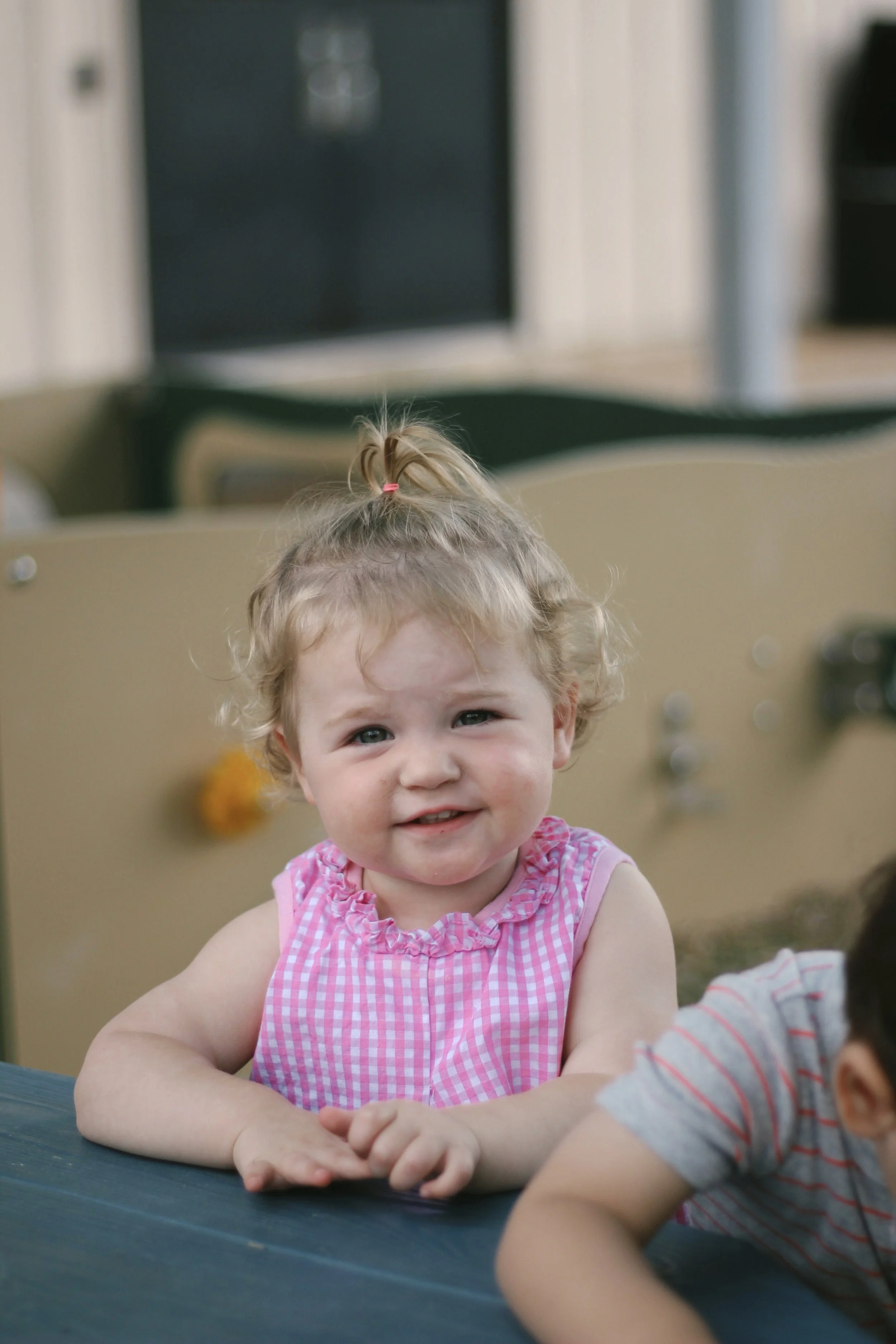 A young girl with curly blonde hair tied in a small ponytail, wearing a pink checked sleeveless dress, sitting at a table outdoors, smiling at the camera.